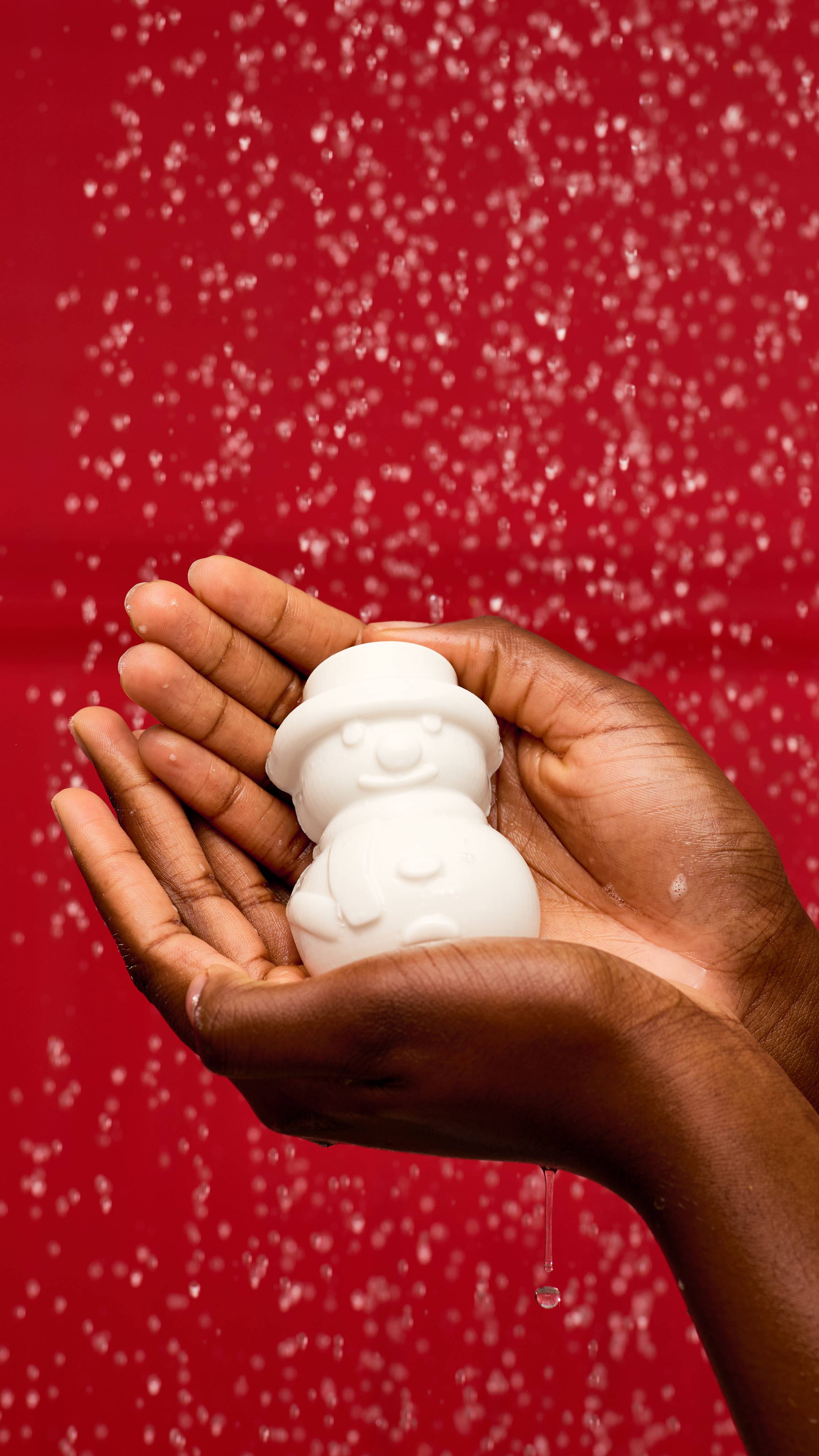 A pair of cupped hands holds a solid, white, snowman-shaped LUSH Snowy shower gummy in the shower. Water droplets are visible in the air, creating a sparkling effect against a vibrant red background.