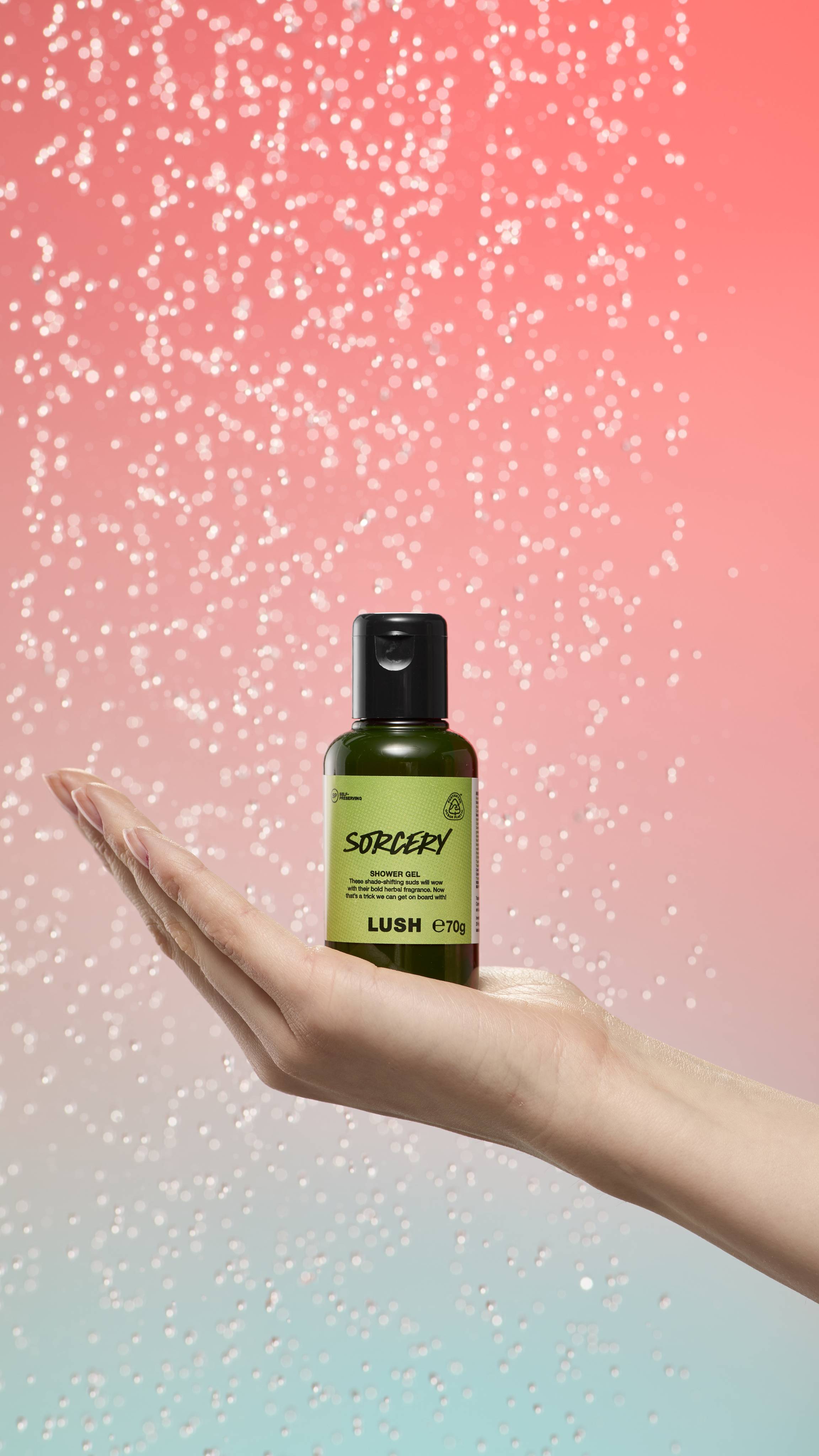 Hand holds a dark green Sorcery shower gel bottle against a shimmering peach-to-blue gradient background. The label is light green with dark green text and the LUSH logo.