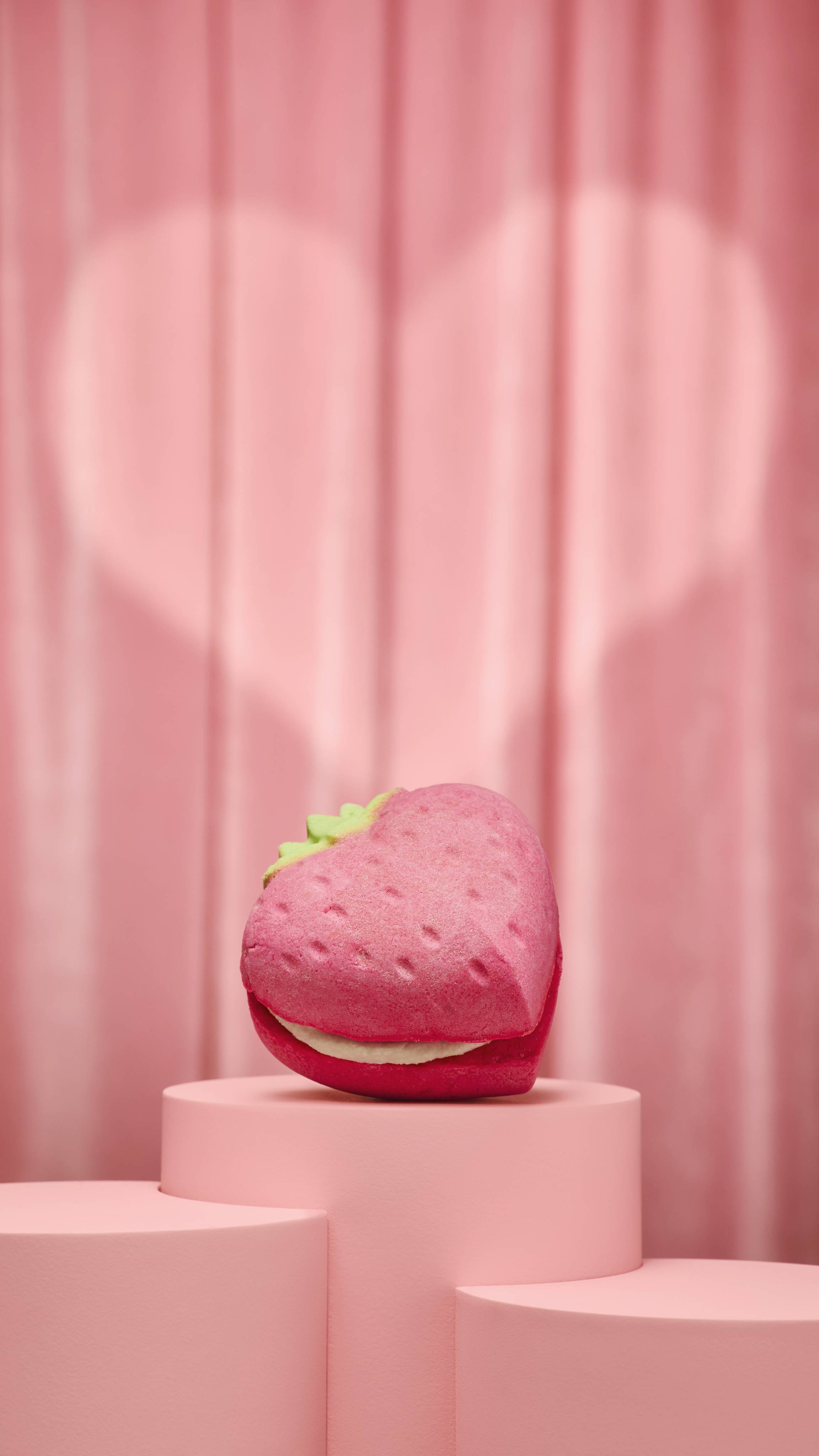 The image shows a heart-shaped bubble bar featuring two pink strawberry halves with textured "seeds" and a green leafy top, sandwiched with a thick layer of white cream. The product sits on a pink podium against a fabric background.