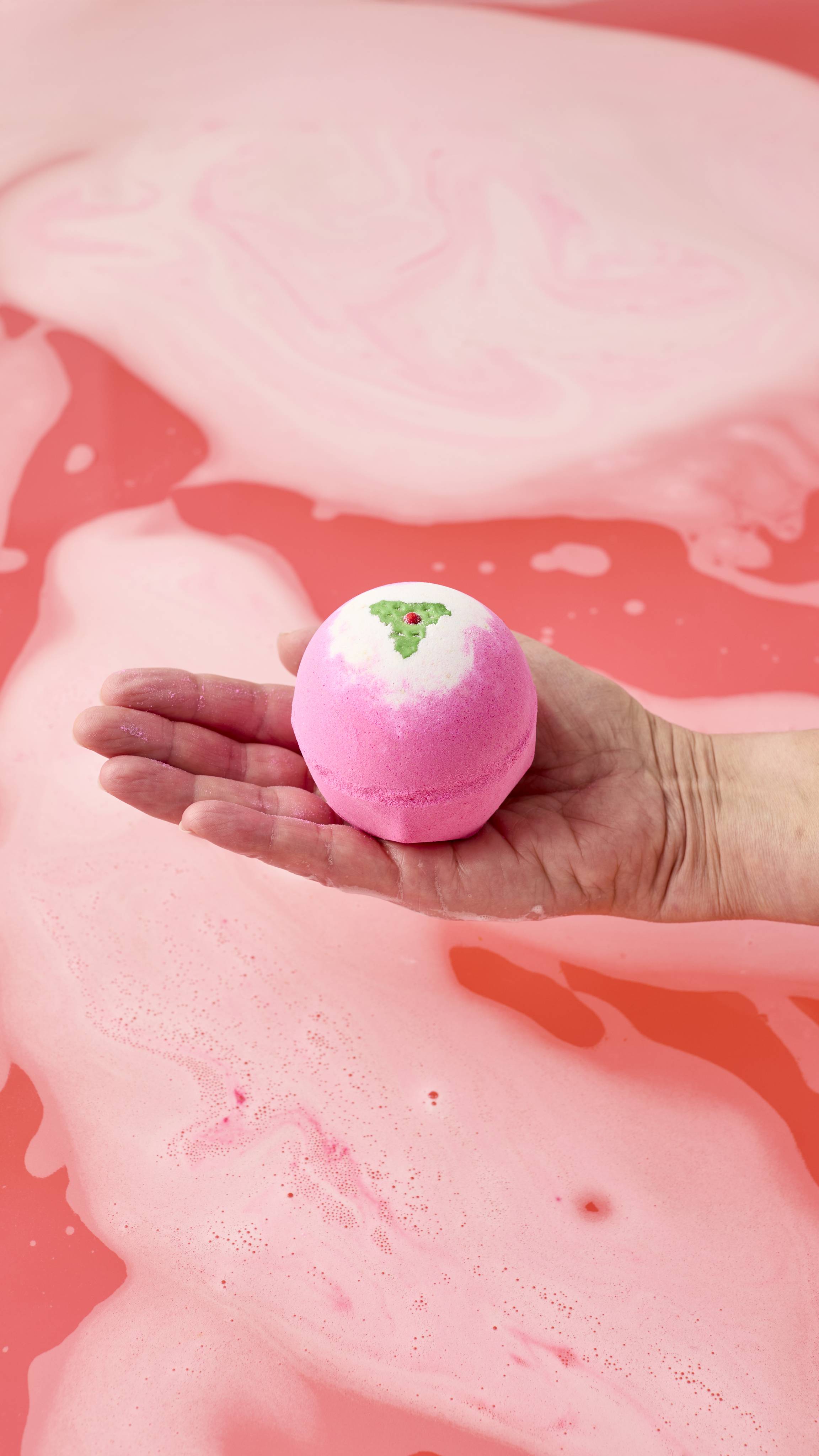The image shows a hand cupping the pink and white Sweet Pudding bath bomb with a small green holly leaf design on top. The product is held just above a vibrant pink bath, with foamy bubbles and swirls.