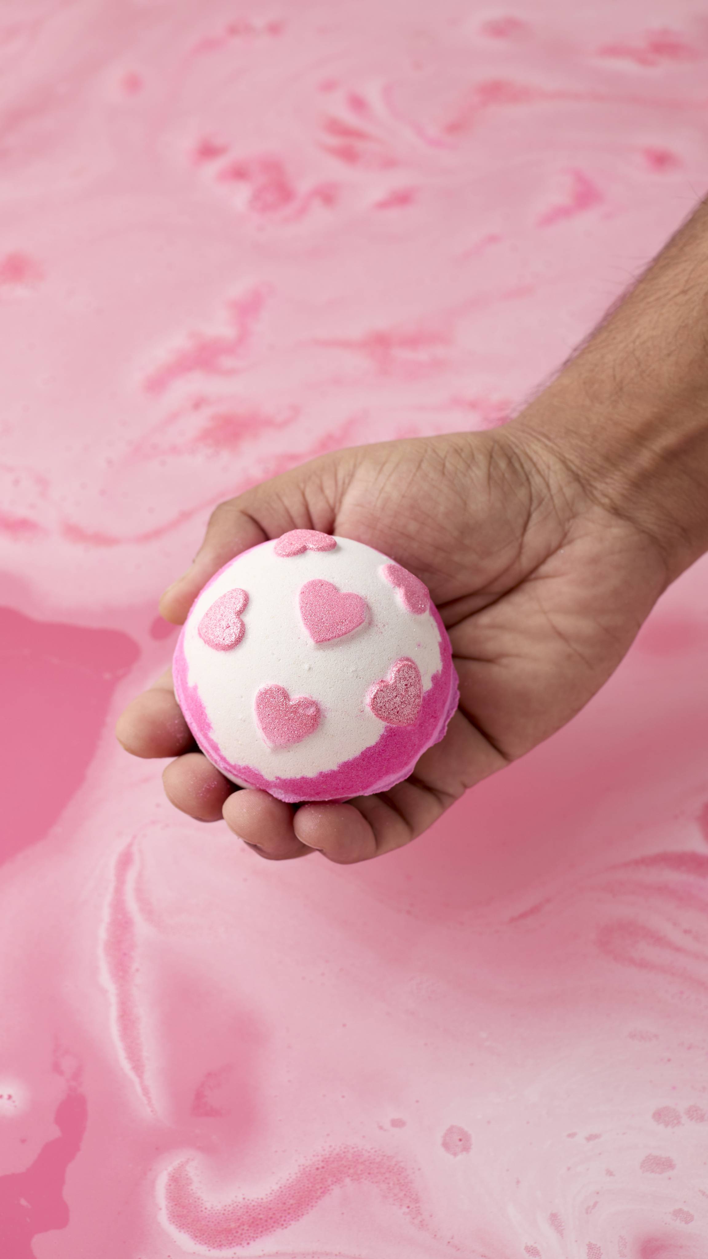 A person's hand holds a round, pink and white Sweetheart bath bomb decorated with small pink hearts above a surface of swirling pink water.