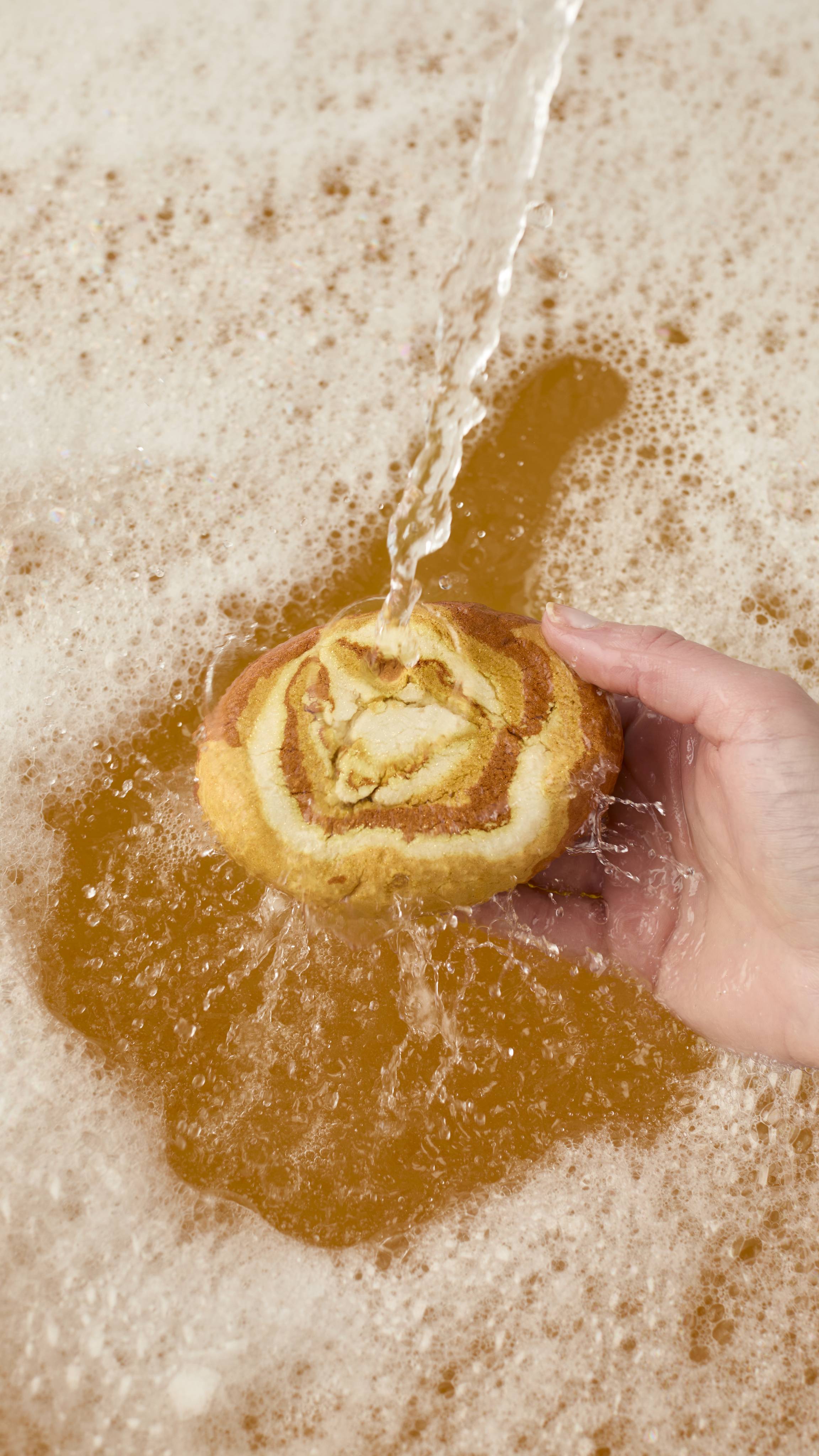 A hand holds the Swirly Sticky Dates bubble bar under a stream of running water, creating a pool of amber-colored water topped with a thick layer of creamy white bubbles.