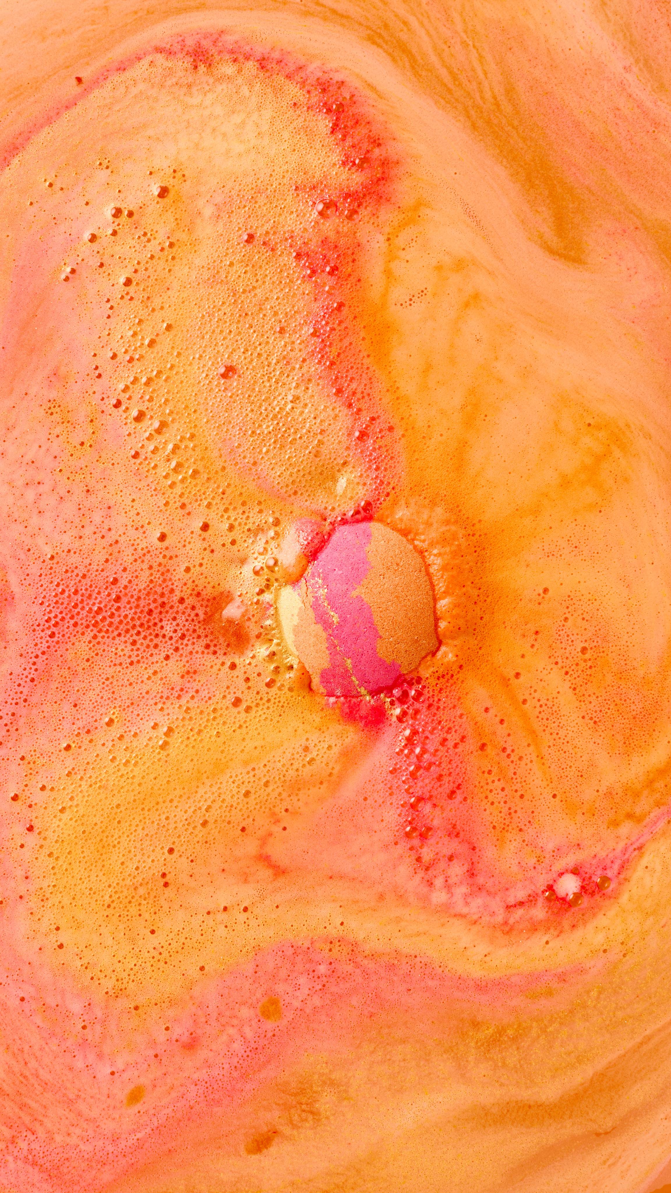 The image shows 'The Alchemist' bath bomb, orange with pink bands, fizzing in the centre of the bath. Vibrant orange, yellow, and red-pink foam swirls outwards, covering the water surface with lively bubbles.