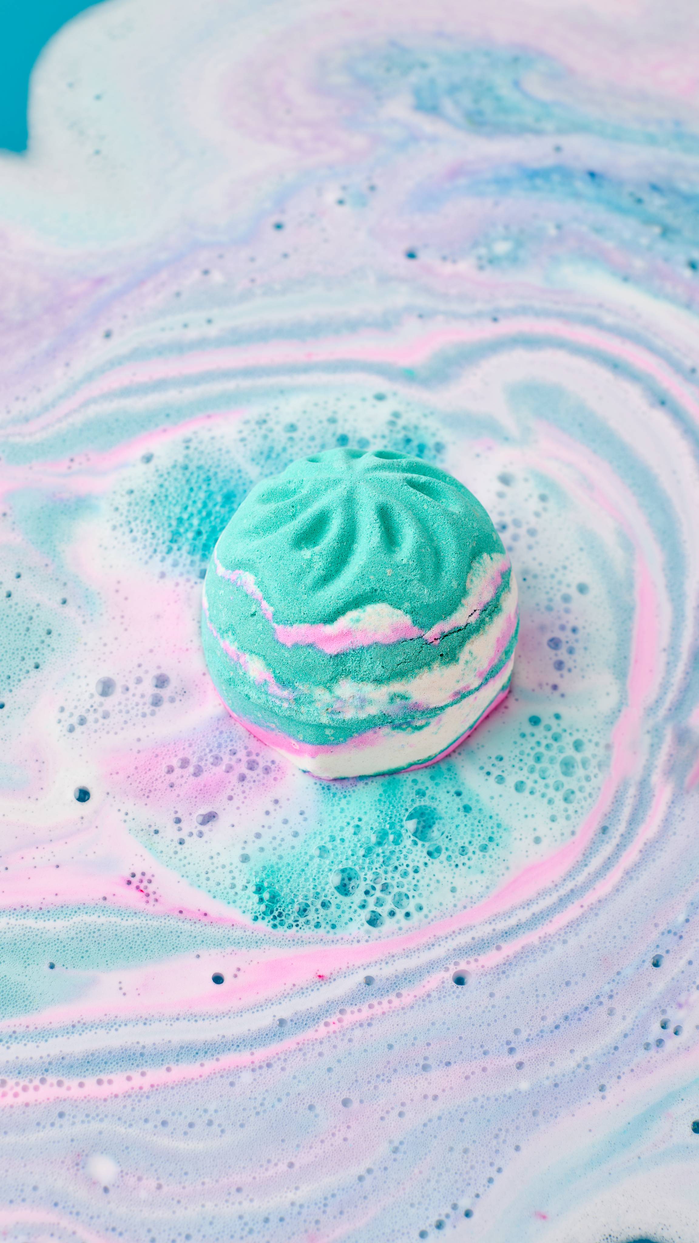 The image shows a marbled 'The Girl In The Whirlwind' bath bomb fizzing in swirling bath water. Vibrant streaks of pink and turquoise bleed from the bomb, creating swirling patterns and white foam across the water.