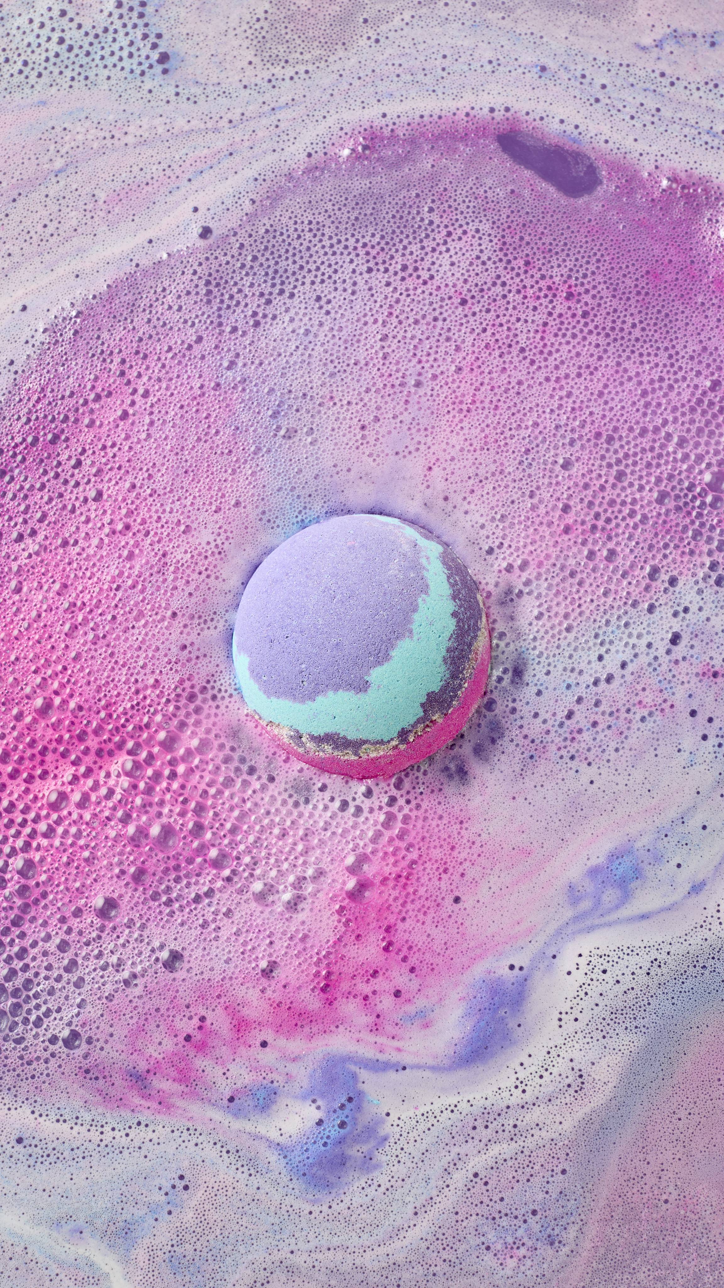 The image shows 'The Magician' bath bomb, banded with purple, light blue, and pink, fizzing in the bath. Vibrant pink foam and bubbles swirl outwards around the bomb, mixing with light purple hues.