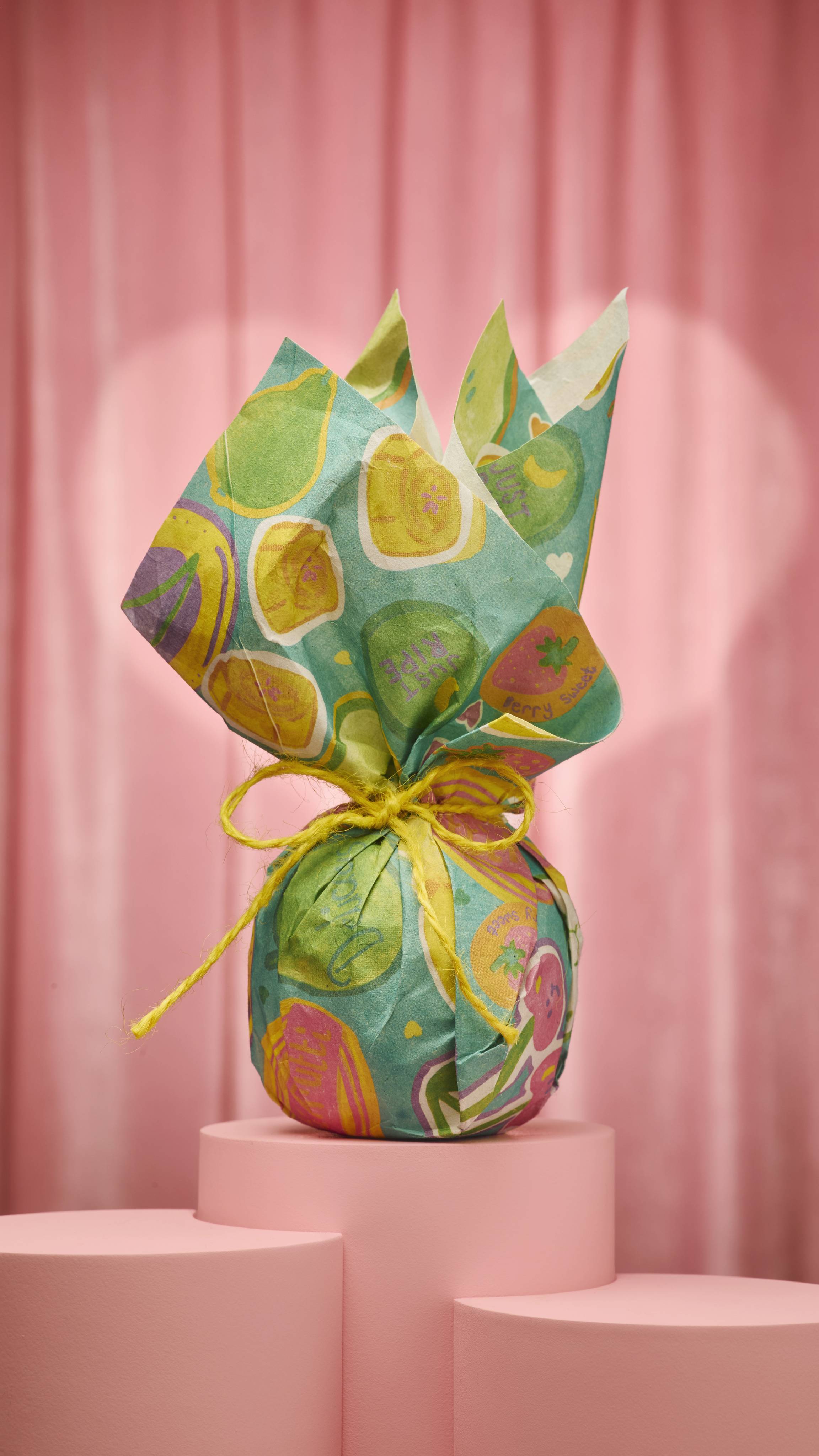 A round gift wrapped in teal Tutti Frutti Lokta paper features yellow lemons, pink strawberries, and "Berry Sweet" text. It is tied at the top with yellow twine against a pink curtain background.