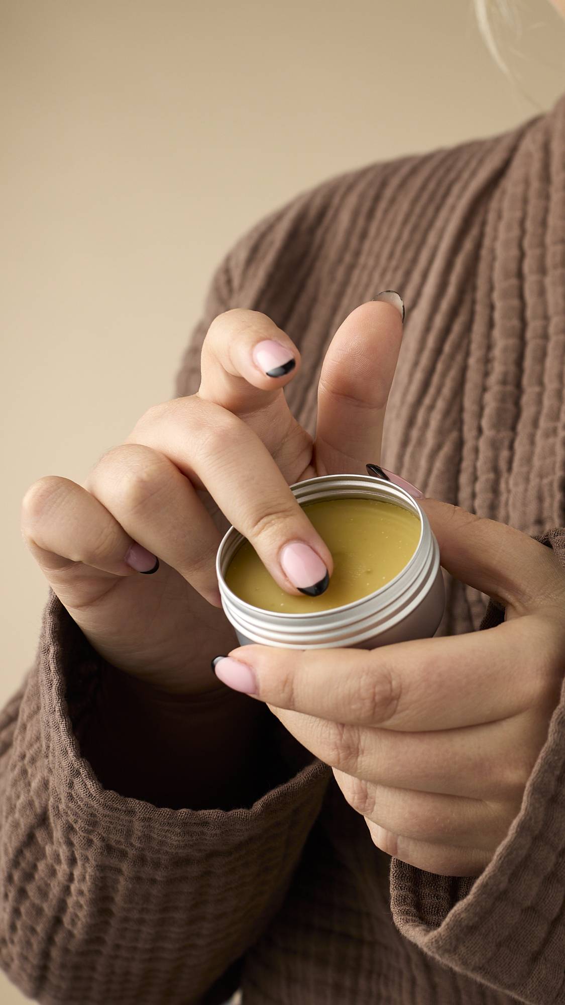 A close-up of a model wearing a brown, textured robe holding a small, round, silver tin of yellow Vision facial serum balm. Their finger is dipped into the product, and they have pink French manicure nails with black tips.