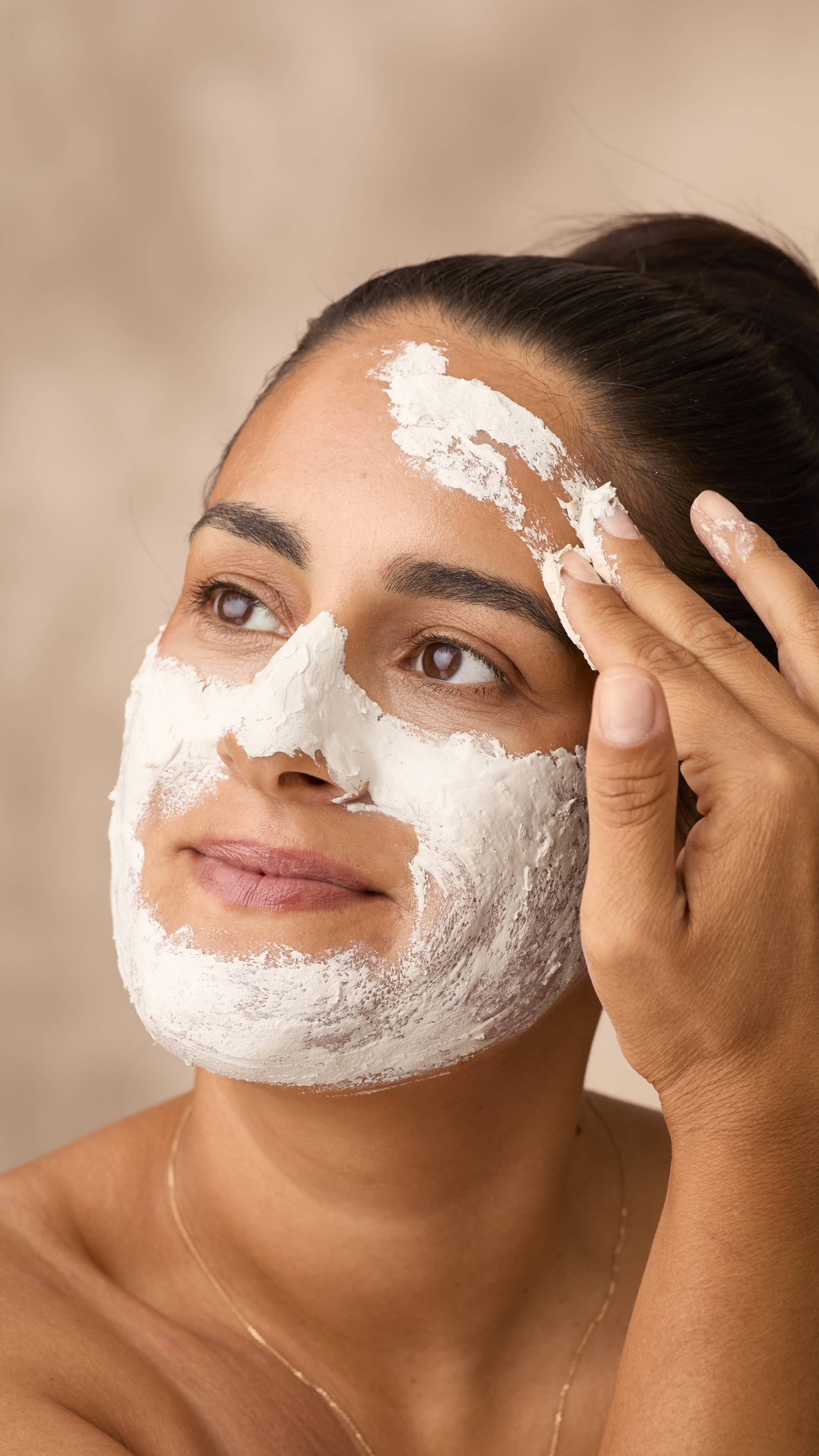 A woman with dark hair tied back looks up and smiles gently while applying a thick layer of Lush Who Runs The World? Ghouls! face mask. The textured paste covers her jaw, nose and cheeks as her fingers smooth it across her forehead.