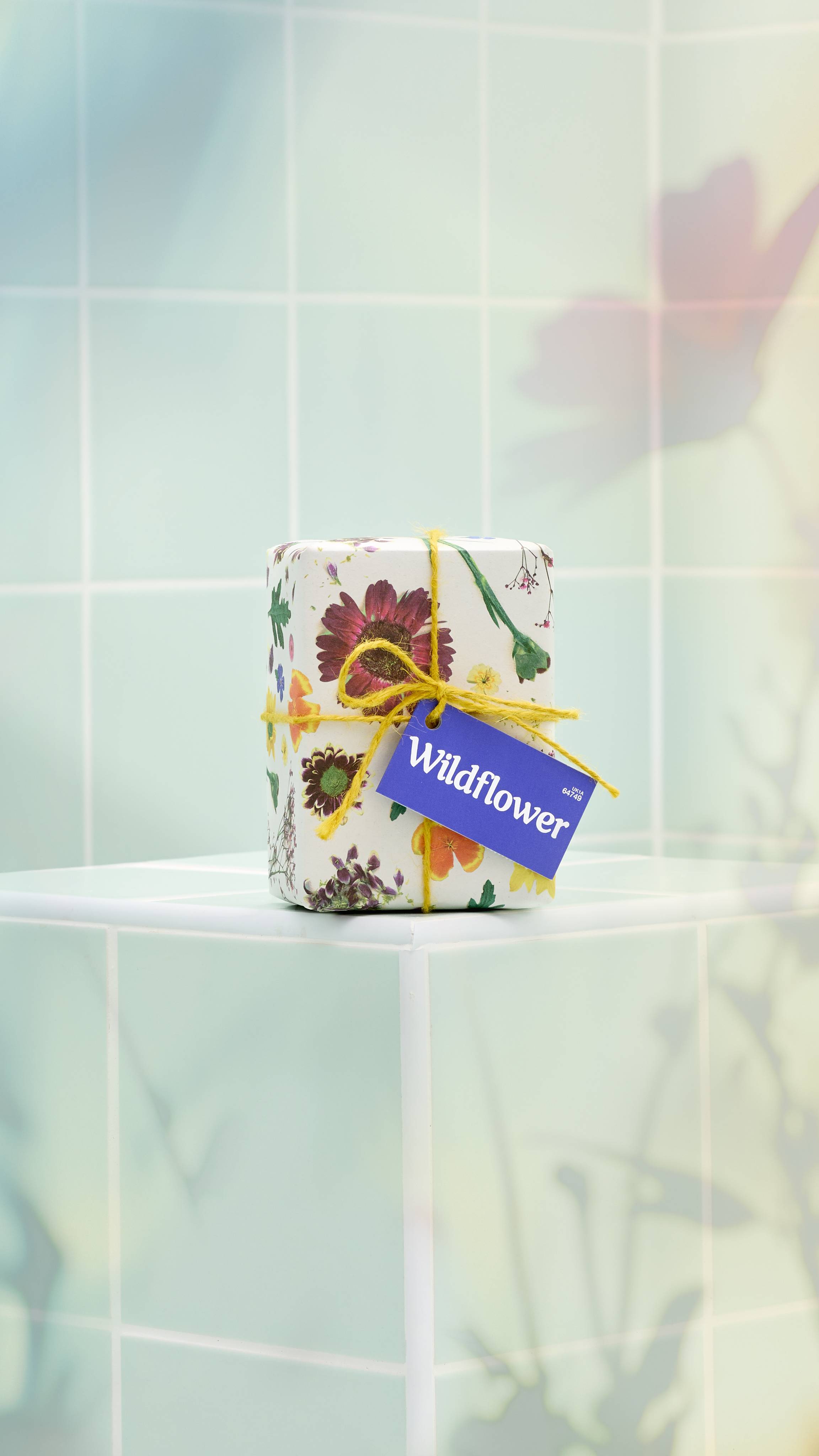 A rectangular "Wildflower" gift wrapped in white paper with a botanical wildflower pattern stands on a pale green tiled ledge. It is tied with yellow twine and features a purple "Wildflower" tag. 