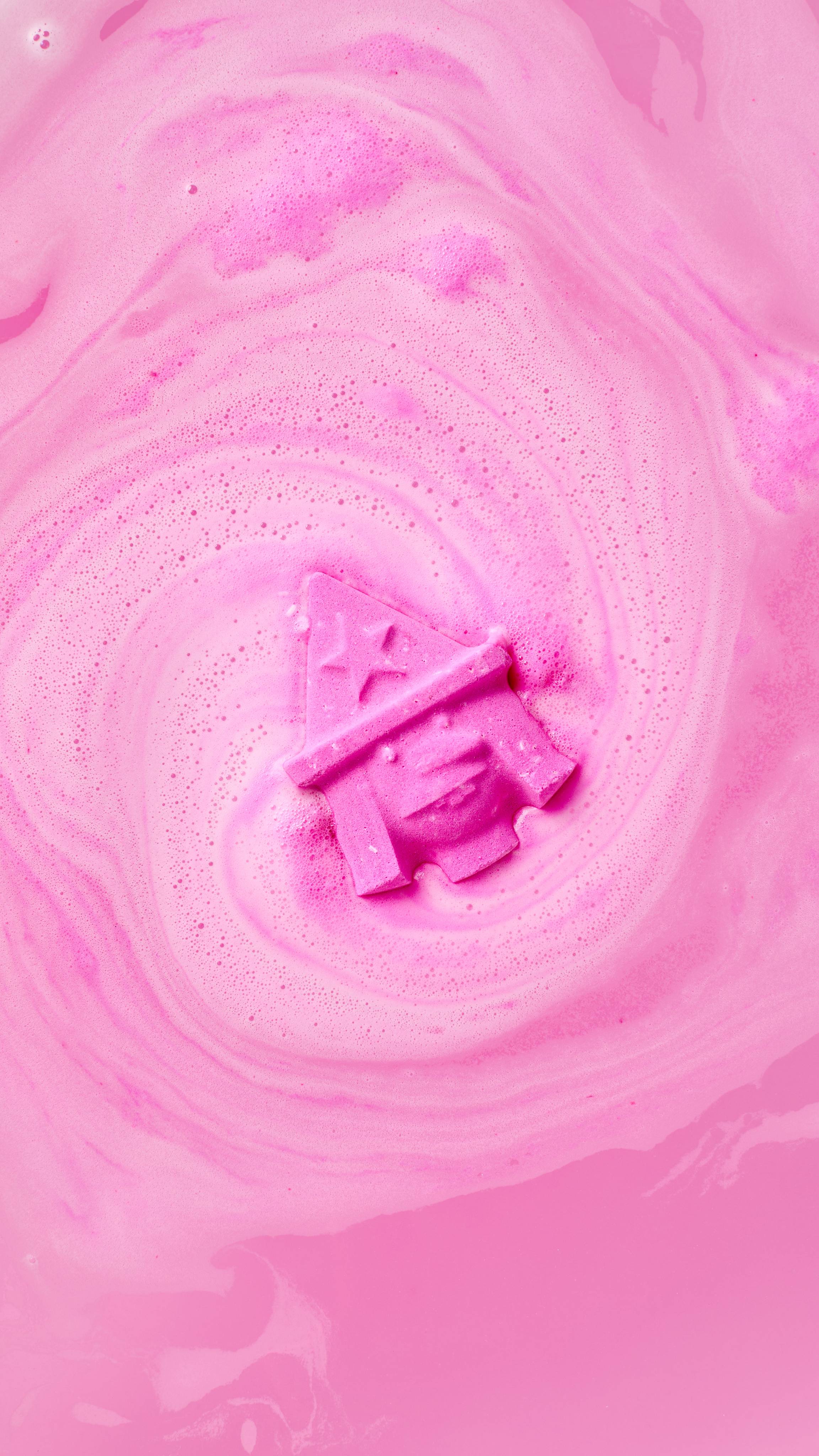 The image shows a vibrant pink 'Witch - Monster Mash Head' bath bomb, fizzing in pink bath water. Rich pink foam swirls outwards in concentric circles around the product, blending with the water.