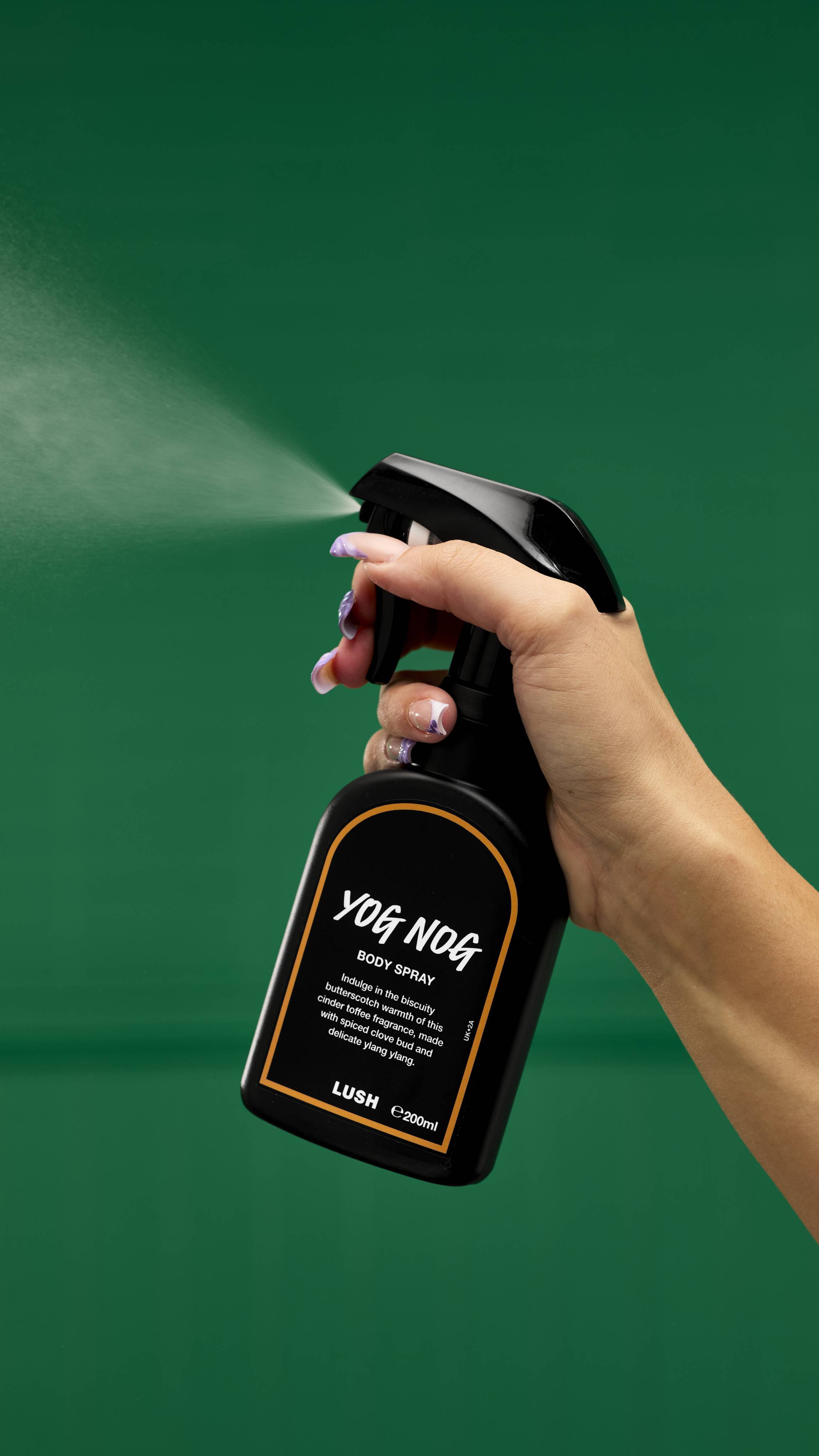A recycled black LUSH body spray bottle with a trigger nozzle is held by a hand with painted nails. The bottle has a gold-rimmed label that reads "Yog Nog Body Spray," and a fine mist of the spray is visible against a vibrant green background.