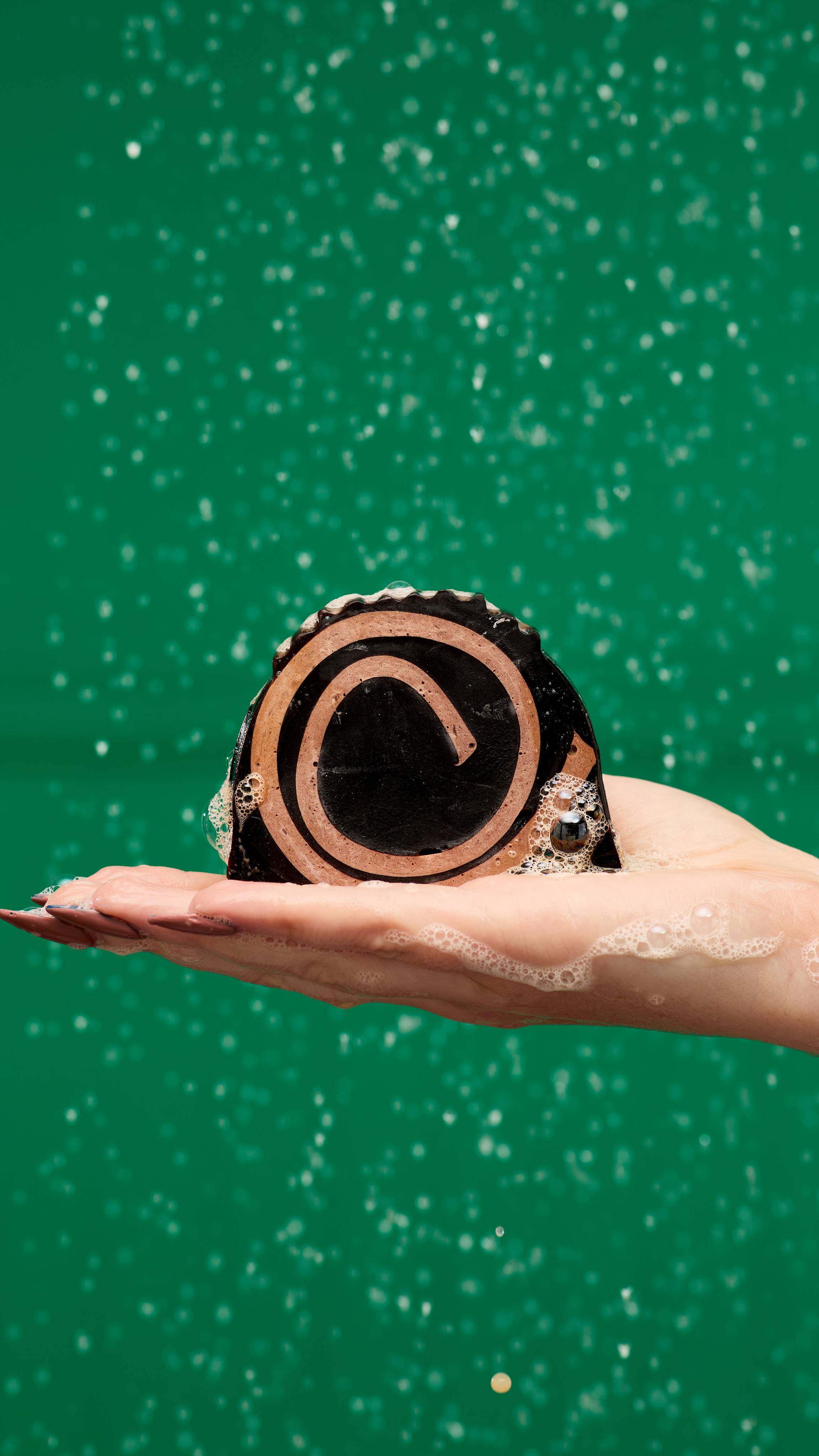 A flat hand holds a slice of Yule Log soap. The soap is dark brown with a light brown swirl design. The soap is creating a lather in the person's hand, and the background is a solid green colour with blurred water droplets.