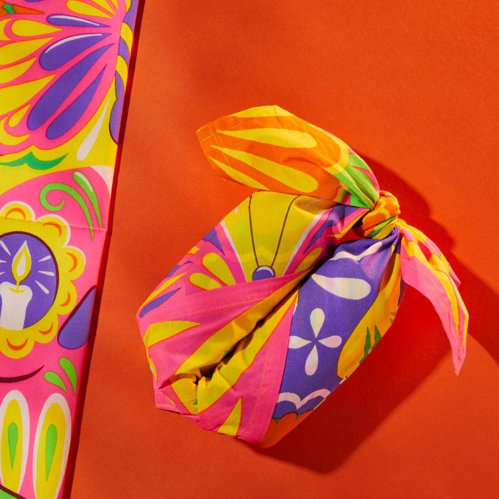 Image shows the Colourful Souls knot wrap tied neatly around a gift on a burnt orange backing with a folded wrap alongside. 
