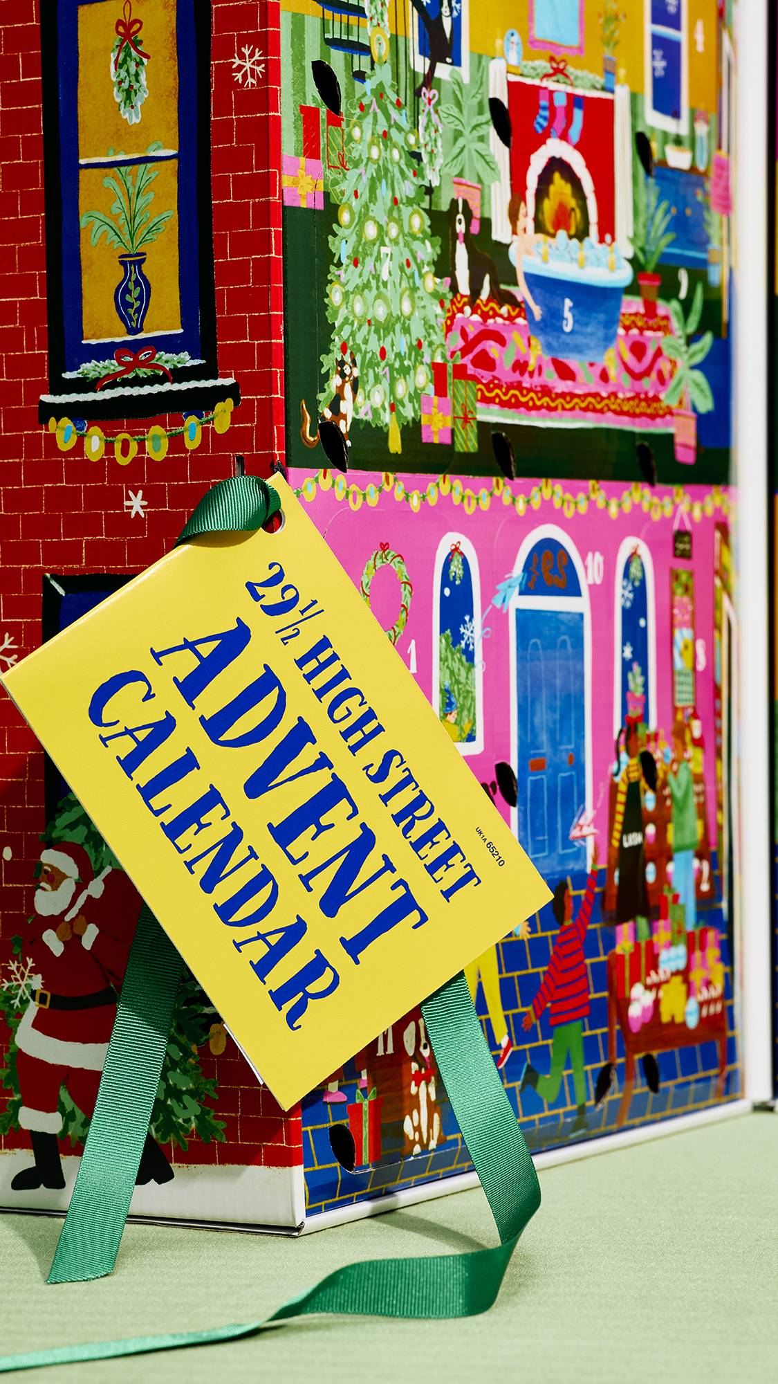 A close-up of a house-themed advent calendar featuring festive illustrated scenes, adorned with a yellow tag tied by a green ribbon that reads "29 ½ High Street Advent Calendar" in blue lettering.