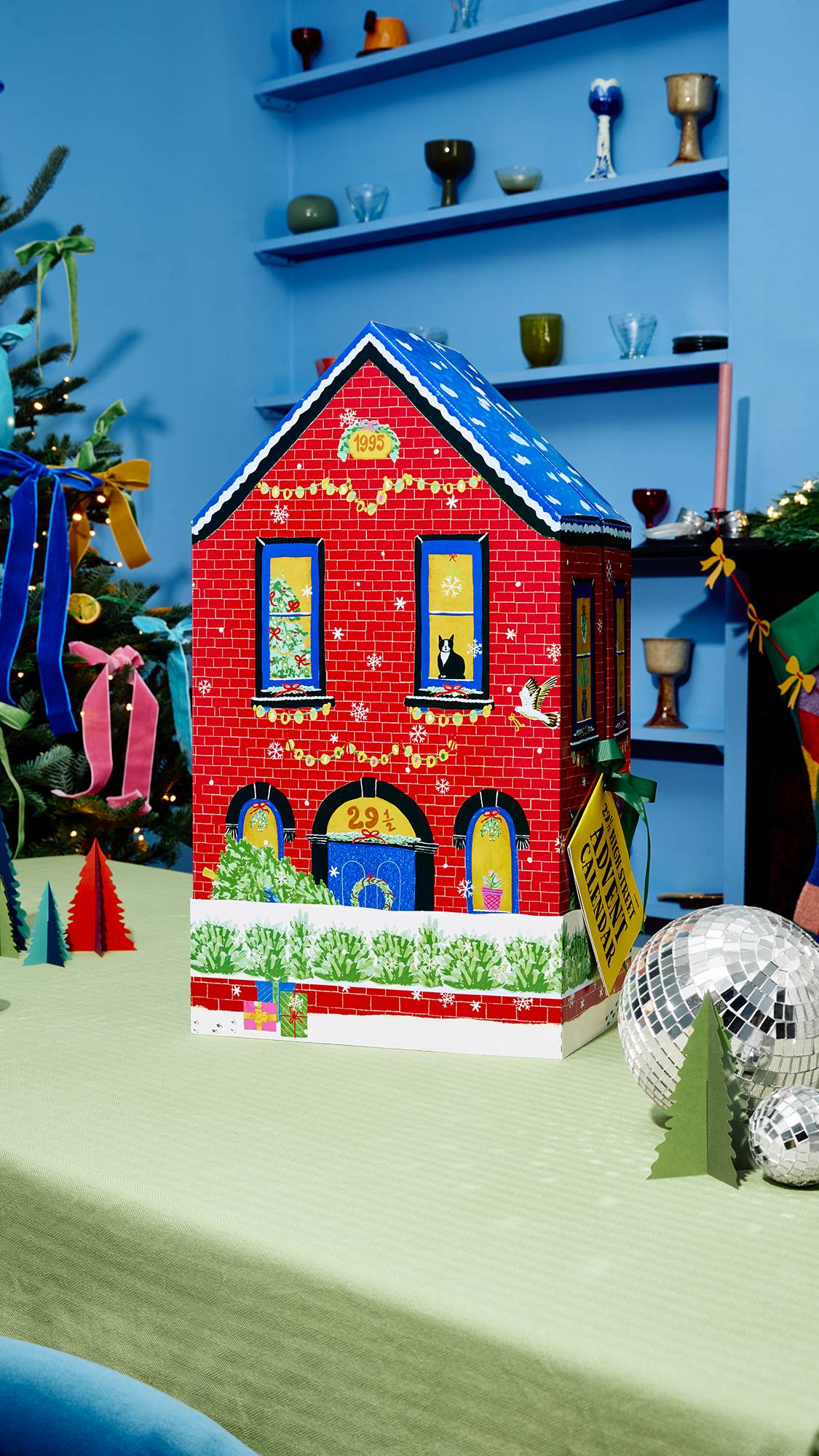 A red brick house-shaped advent calendar with a blue snow-dusted roof stands on a green table, decorated with Christmas lights, a black cat in the window, and a yellow tag attached, set beside silver disco balls.