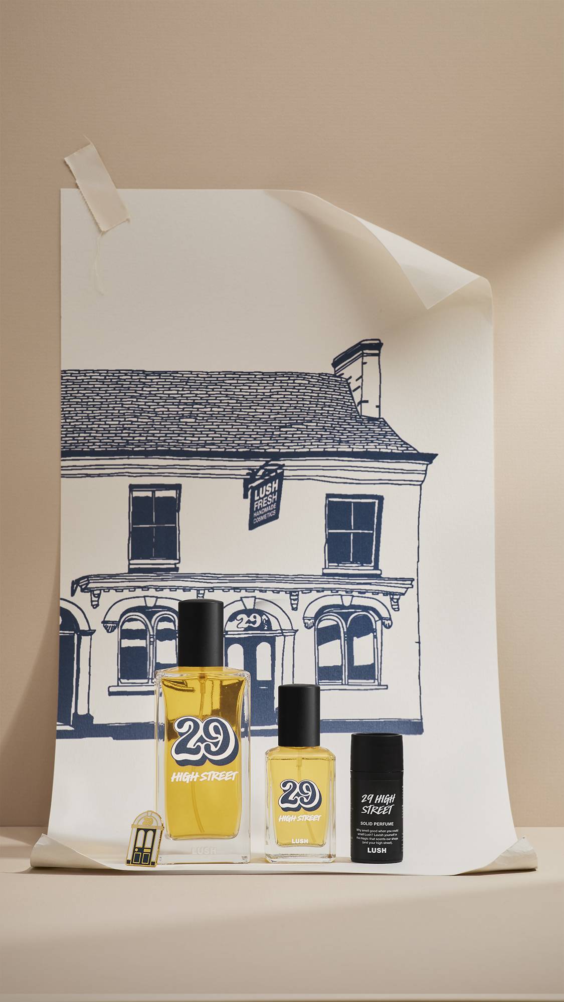 Four products from the Lush '29 High Street' range—two glass perfume bottles, a metal pin badge and a solid perfume tin—stand in front of a navy-blue line drawing of the original Lush shop on a large poster. 
