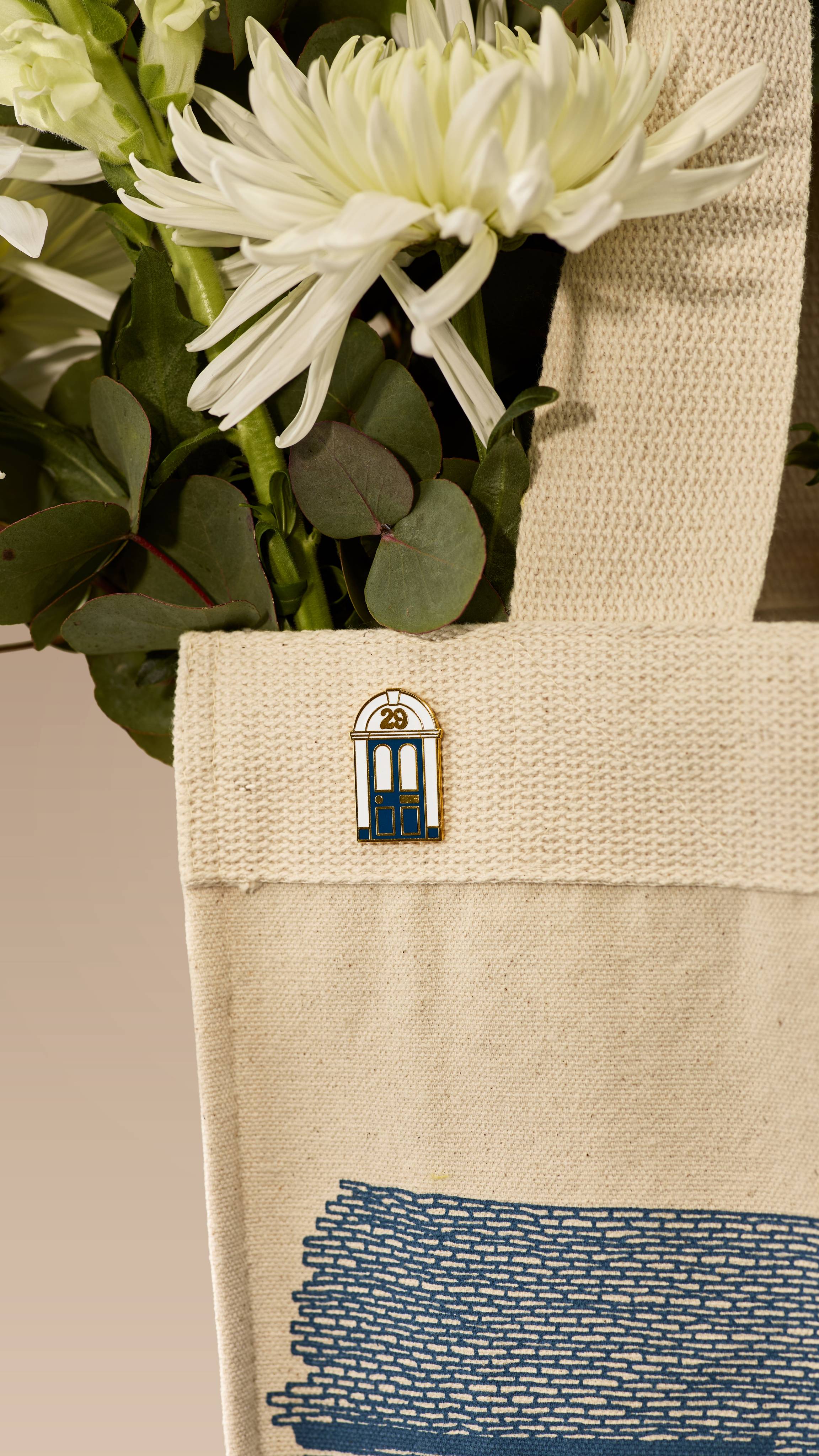 A close-up shot of a cream canvas tote bag containing white flowers and eucalyptus. A small enamel pin, shaped like a navy-blue doorway with the number '29' at the top, is attached to the upper edge of the bag.