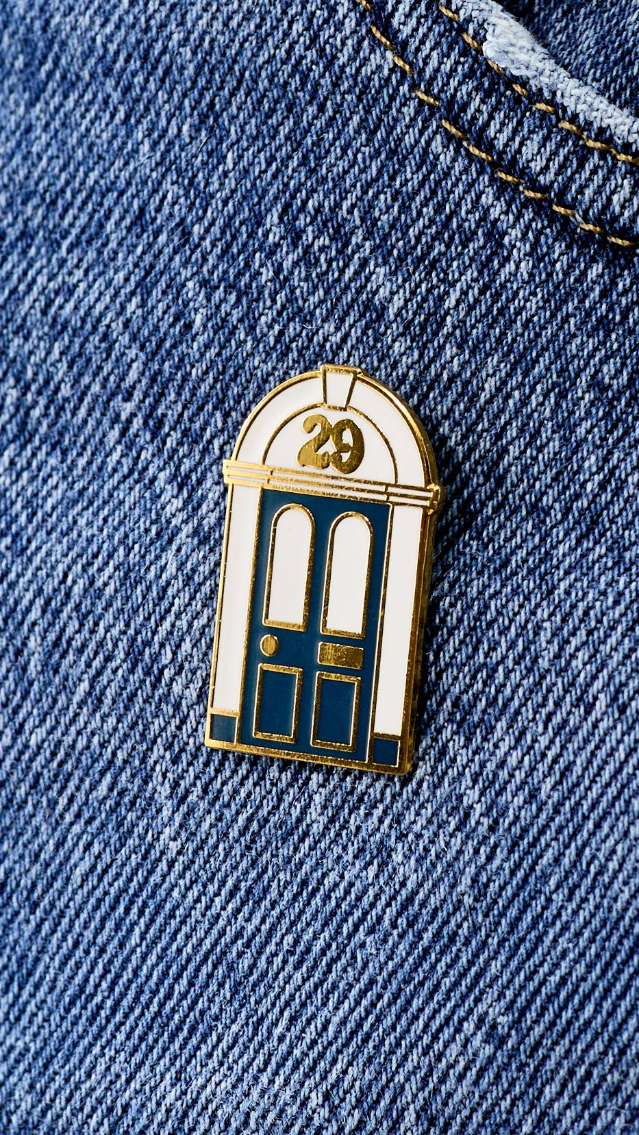 An enamel pin shaped like a traditional doorway with a navy-blue door and white surround is pinned to blue denim fabric. The number '29' is featured in gold at the top of the arched frame. 