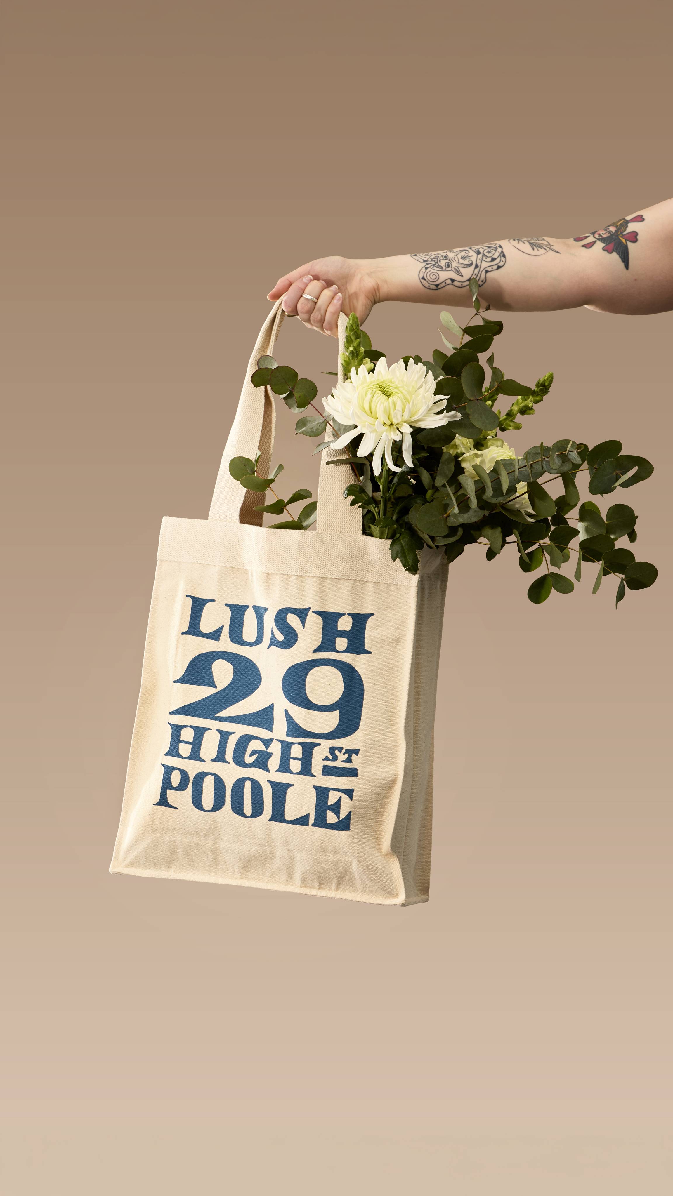 A tattooed arm holds up a cream canvas tote bag filled with white flowers and eucalyptus. The bag features large navy-blue text reading 'LUSH 29 HIGH ST POOLE' in a vintage-style font, set against a neutral beige background.
