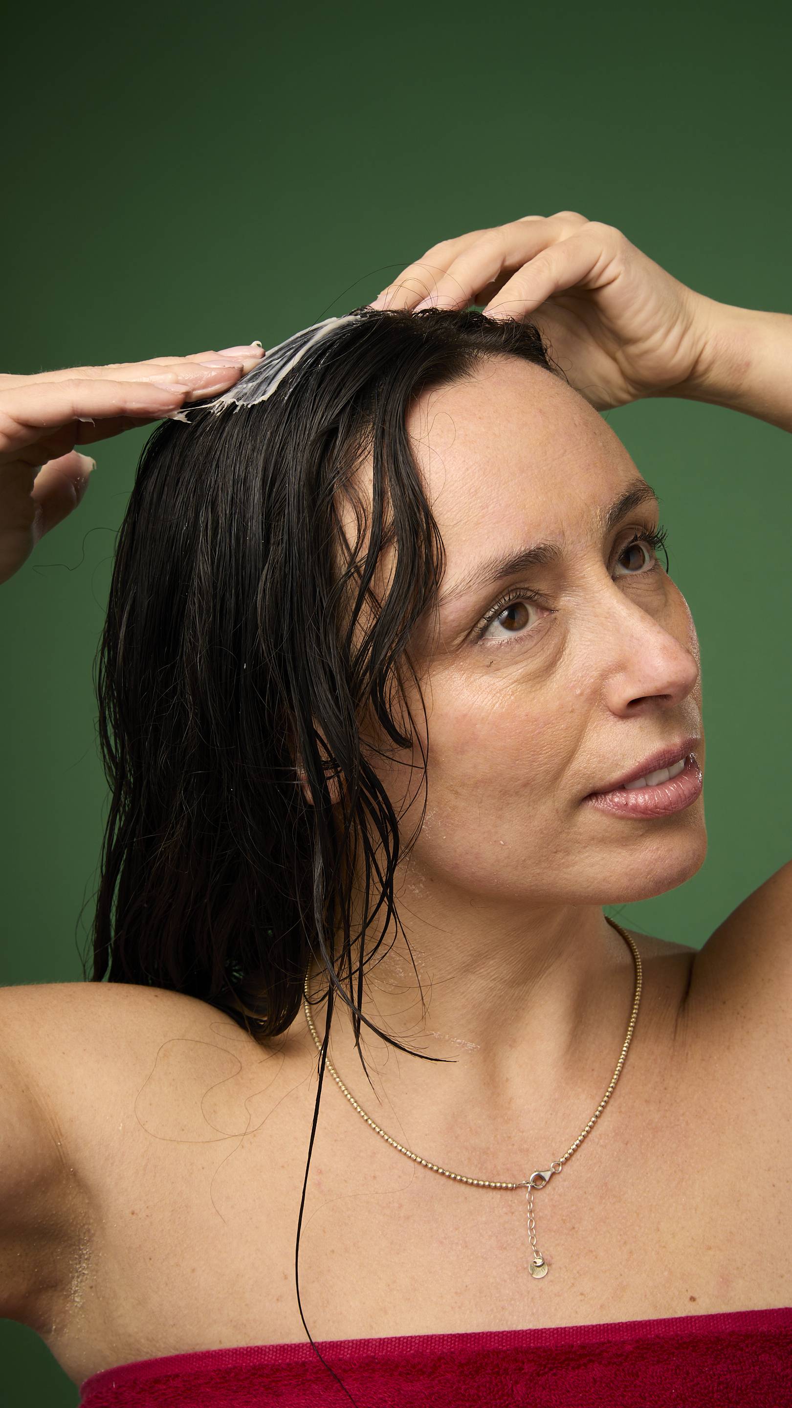 A model with long, wet, dark hair is shown from the chest up against a solid green background. They are applying the Lush Feast of Fools hair treatment directly to their scalp.