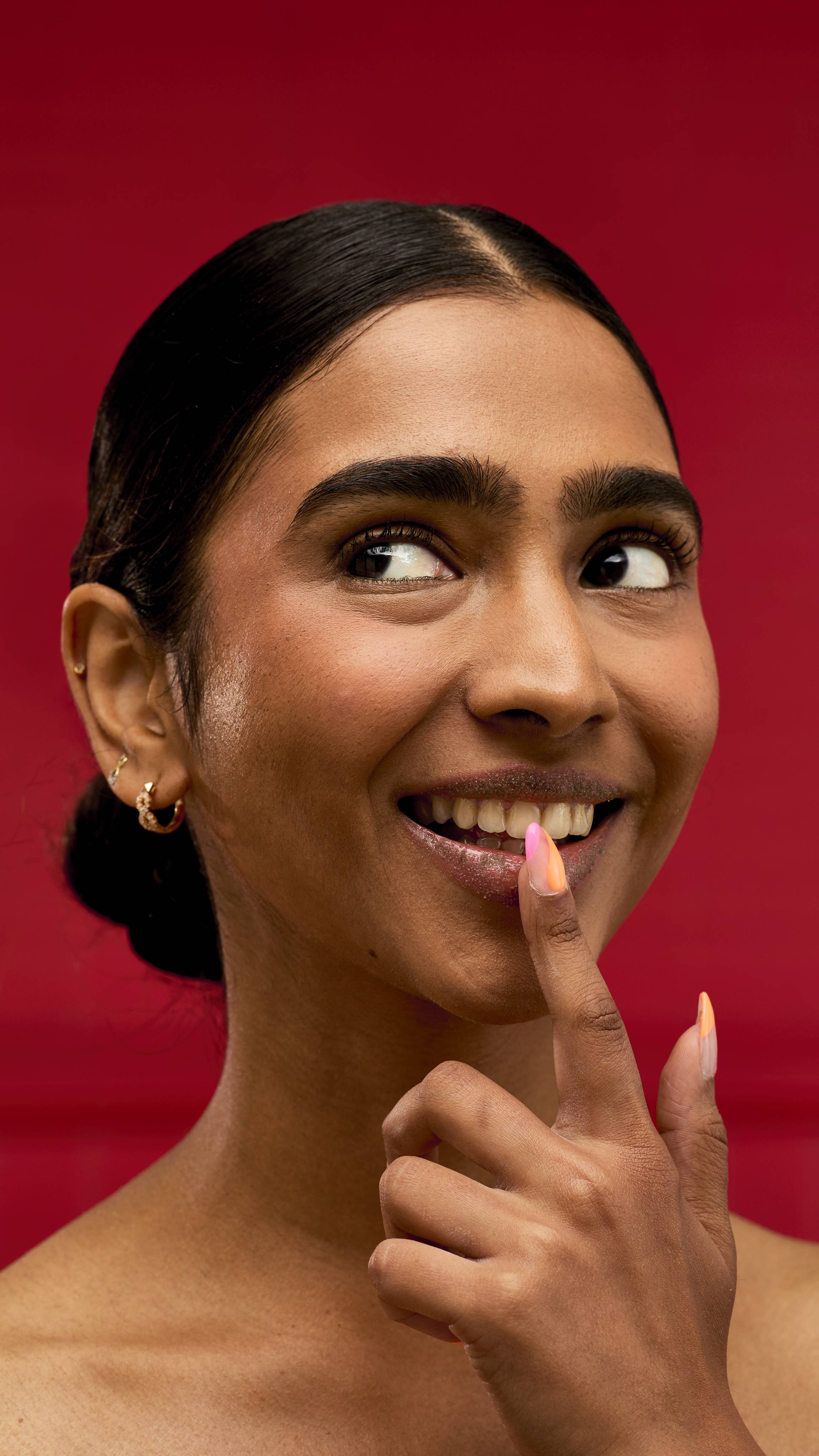 A person smiles playfully while touching their lip with a finger that has a colourful ombré manicure. Their lips are coated in a subtle sheen, suggesting a recently applied LUSH After Dinner Mint lip scrub. The background is a vibrant red.