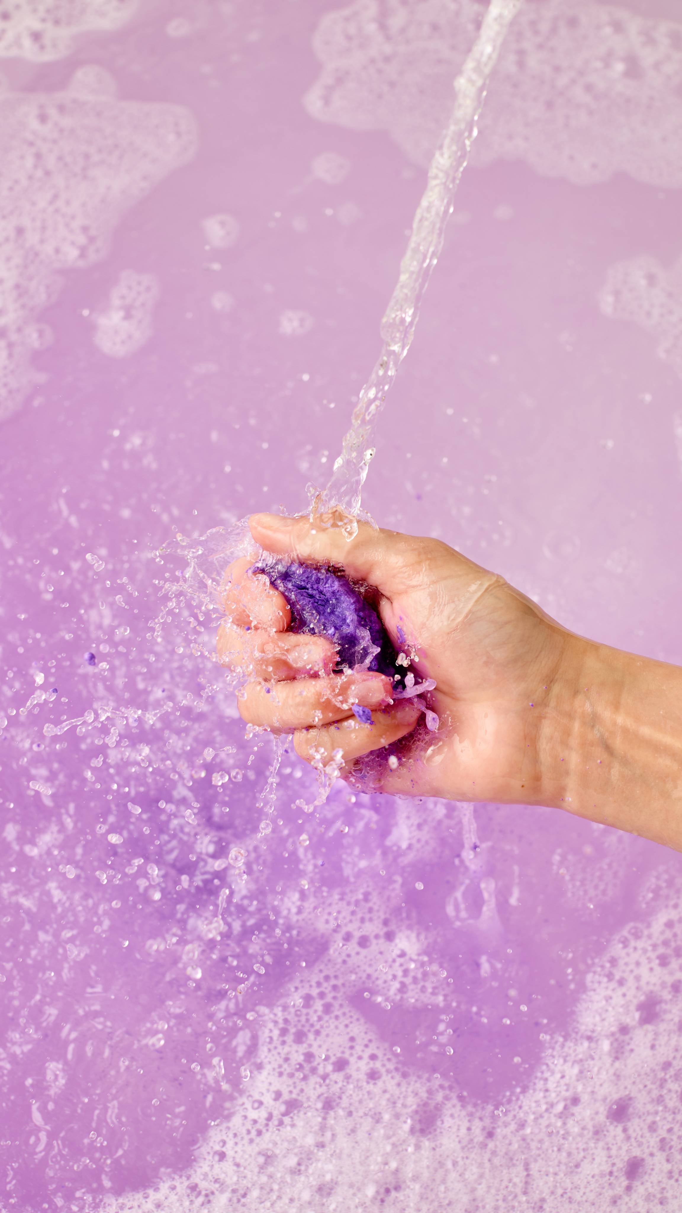 The image shows a light-toned hand vigorously squeezing and crumbling a vibrant purple 'Alien' bubble bar under a strong stream of water. Abundant purple foam and splashes explode across the bath.
