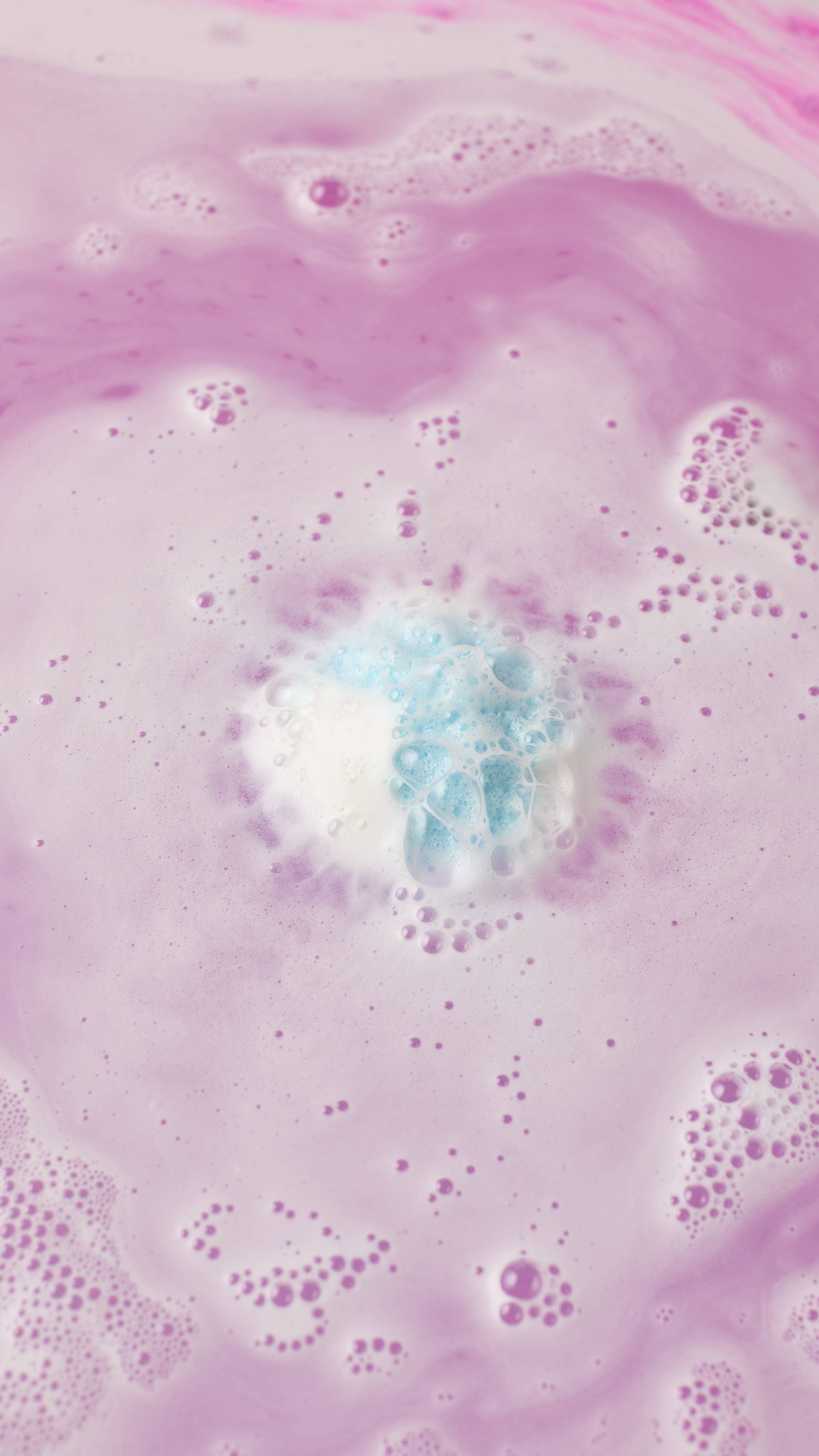 An aerial view shows a small, blue-and-white "Atom Heart Mother" bath bomb dissolving in water. It creates a swirling pattern of pale purple and blue foam and bubbles that spread across the surface of the pink water.