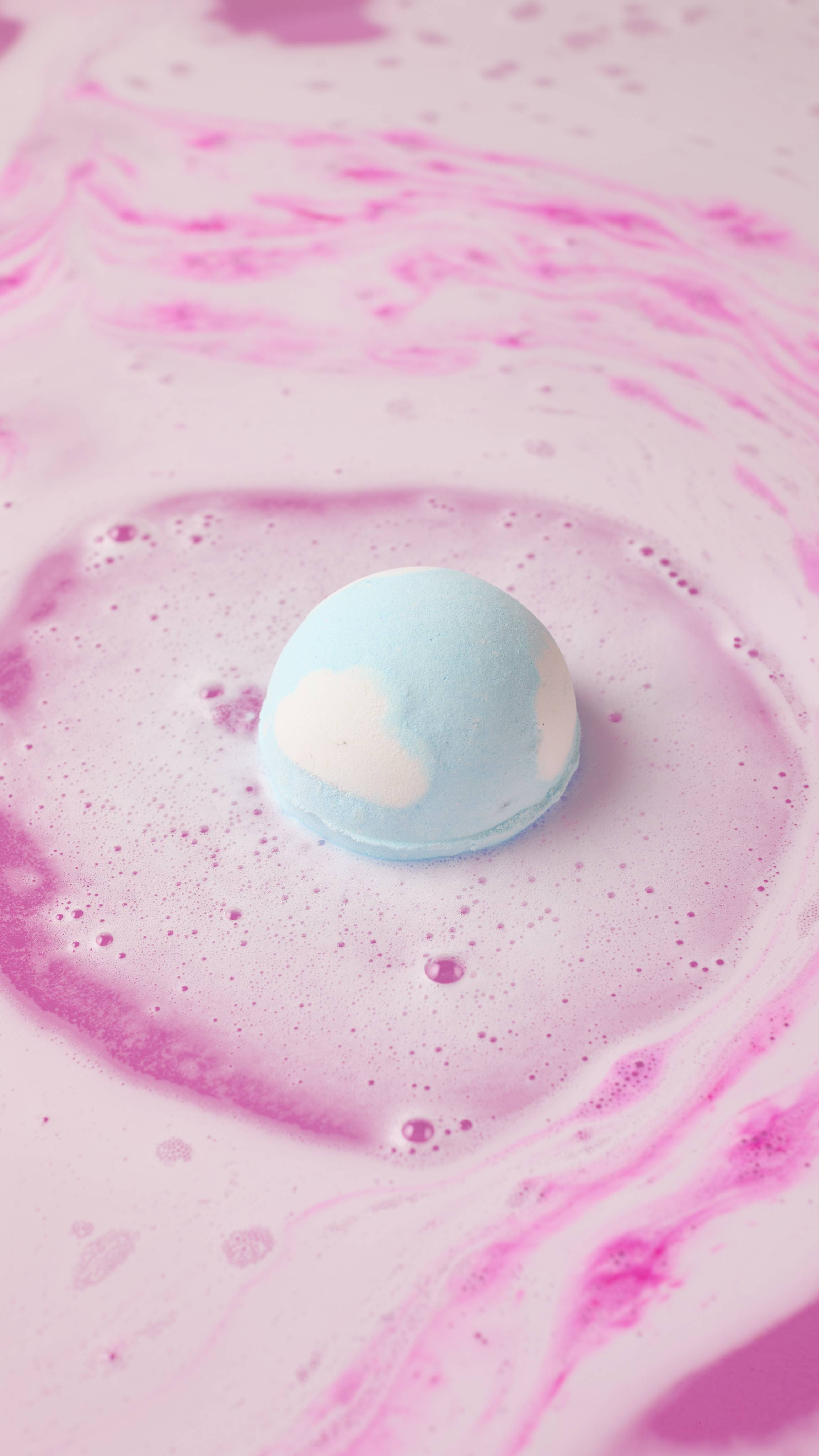An aerial view shows a small, blue-and-white "Atom Heart Mother" bath bomb dissolving in water. It creates a swirling pattern of pale purple and blue foam and bubbles that spread across the surface of the pink water.