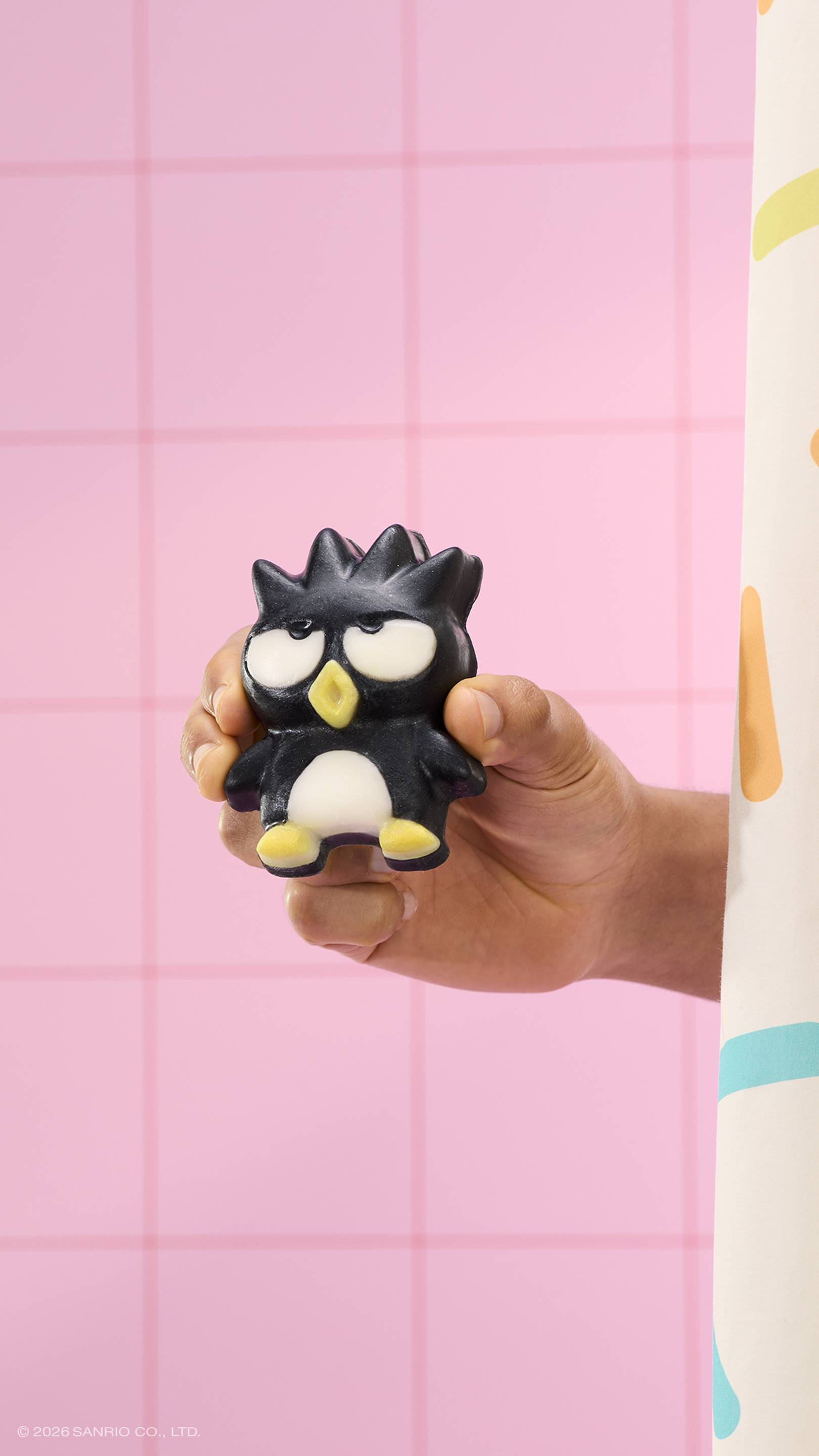 A hand holds a black penguin-shaped soap with spiky hair and a grumpy expression against a pink tiled wall. A sprinkle-patterned shower curtain is visible on the right. The text '© 2026 SANRIO CO., LTD.' is at the bottom.