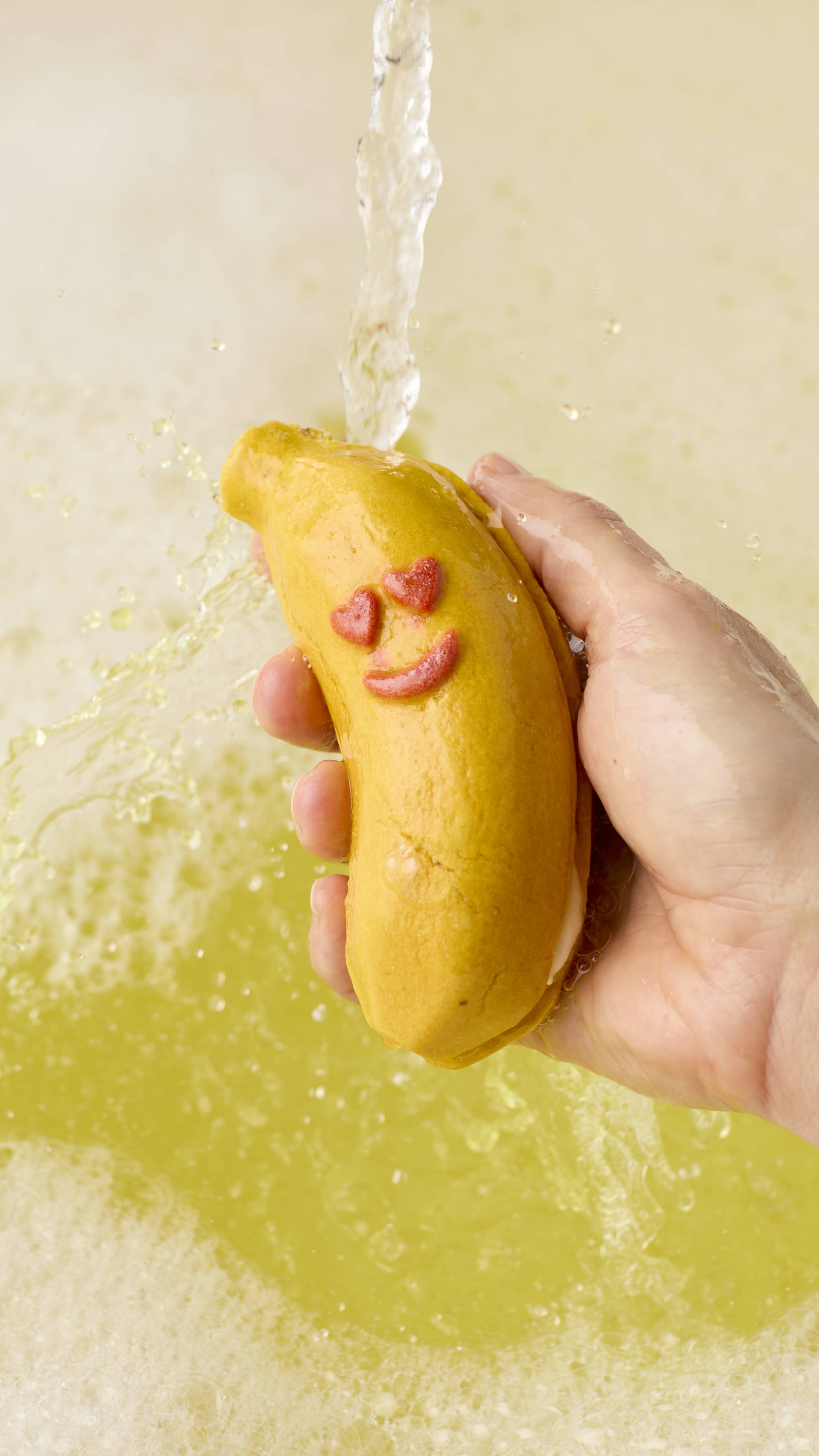 The image shows a hand holding the banana-shaped bubble bar under a stream of running water. The water splashes against the bar, creating pale yellow bubbles and foam in the bath below.