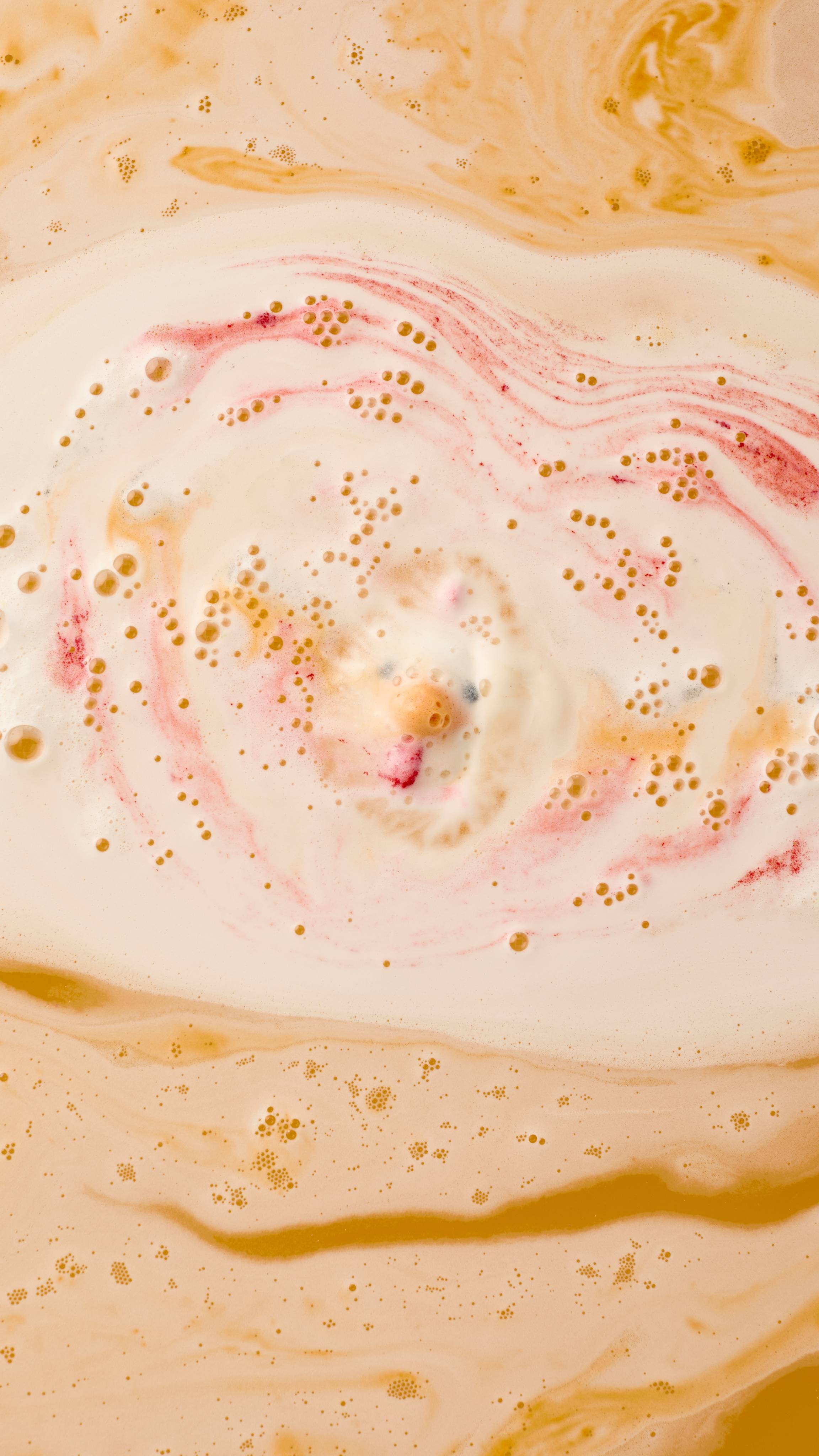 The Big Dilemma bath bomb dissolves in water, creating a center of creamy white foam surrounded by expanding swirls of deep orange and bright pink. Tiny bubbles gather on the surface of the marbled water.