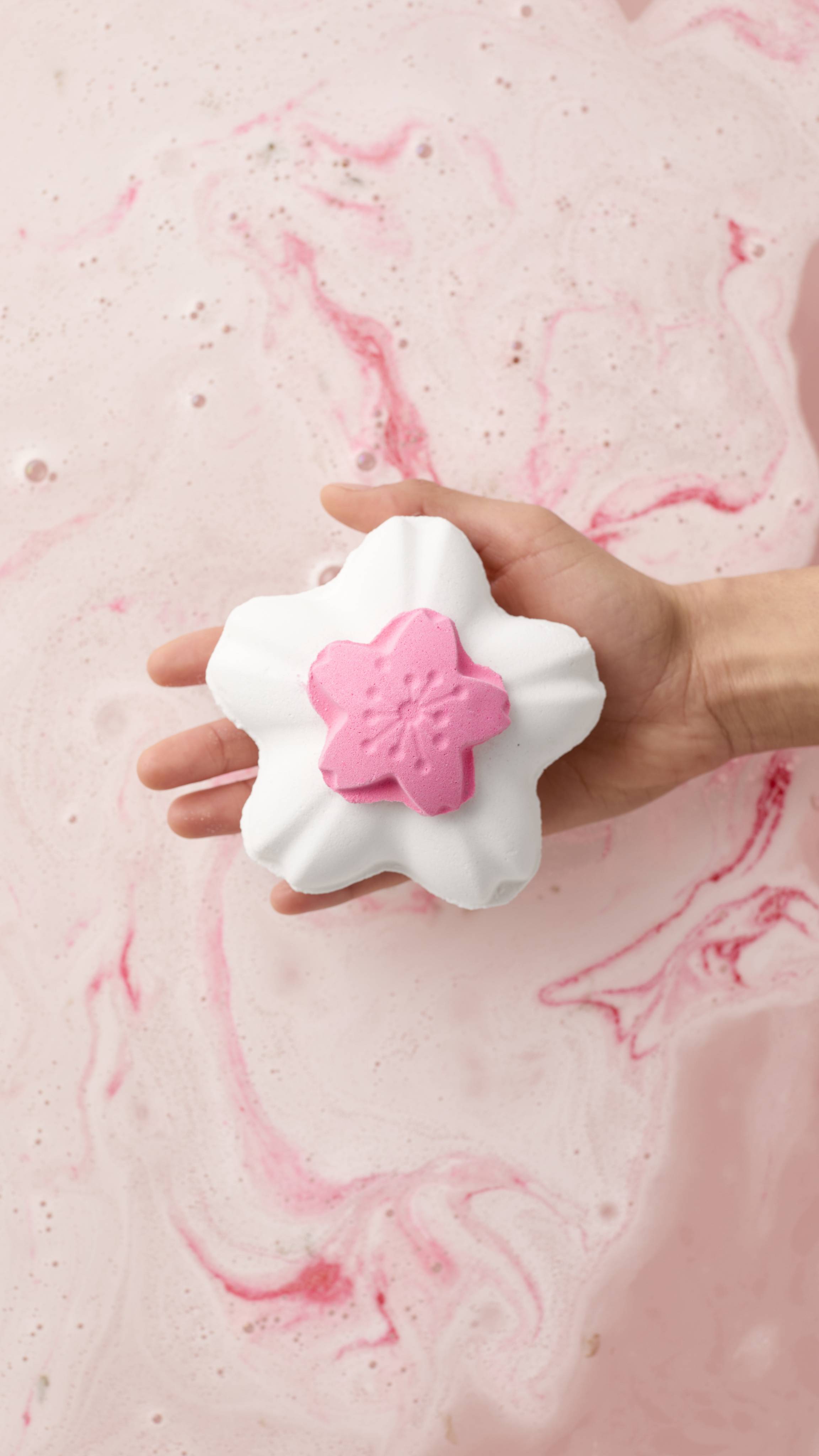 A model's hand holds the "Blooming Cherry Blossoms" bath bomb with a pink floral centre. It is held over bathwater filled with swirls of pale pink foam and vibrant pink patterns. 