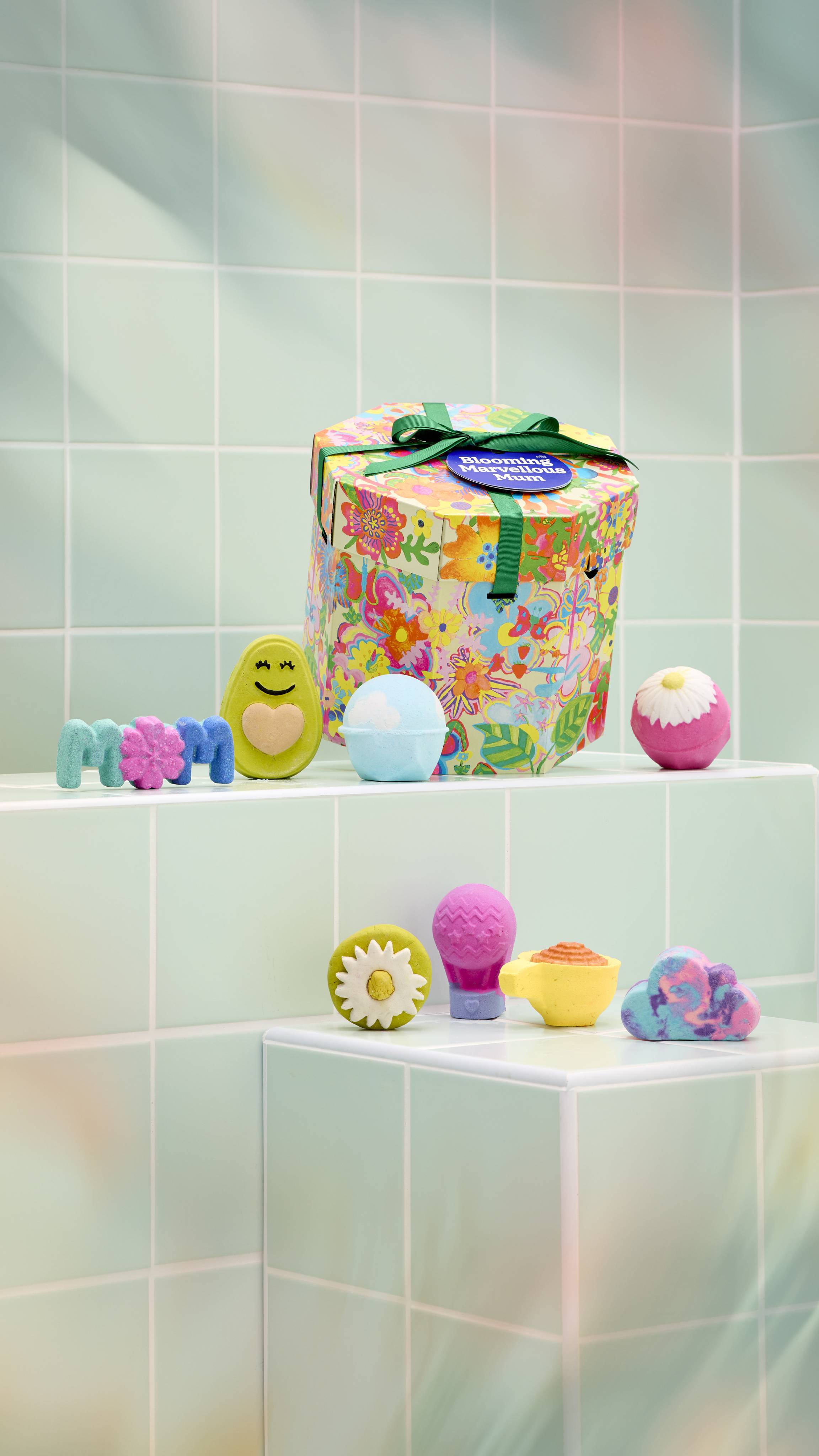 A vibrant, hexagonal "BloomingMarvellous Mum" gift box with a floral pattern sits on a tiled ledge. Arranged in front are various bath products, including five bath bombs and three bubble bars.