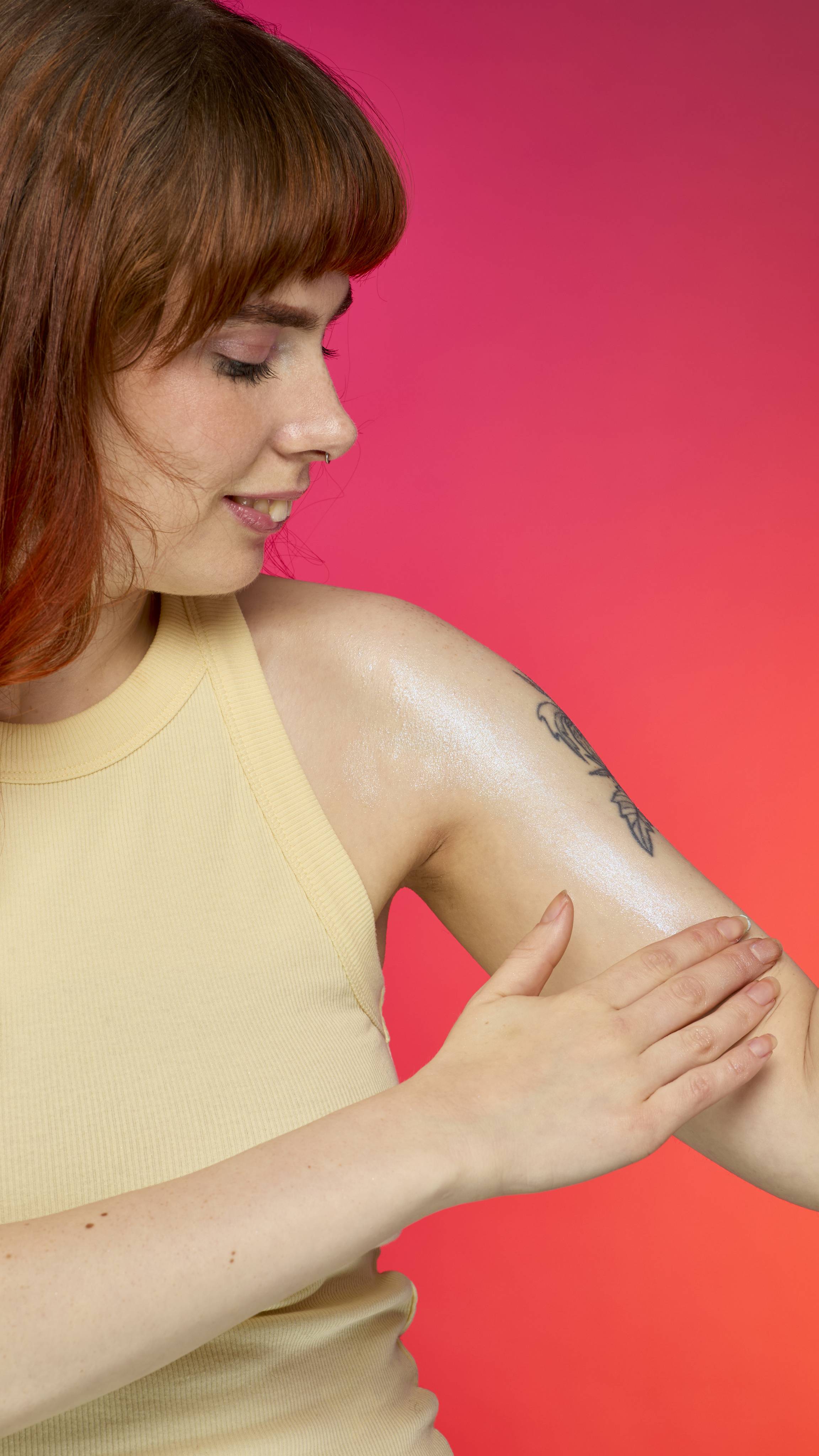 A person with red hair applies the faceted, pyramid-shaped Blue glitterbug body bar to their tattooed arm. The skin shimmers with plastic free glitter against a magenta and red background.