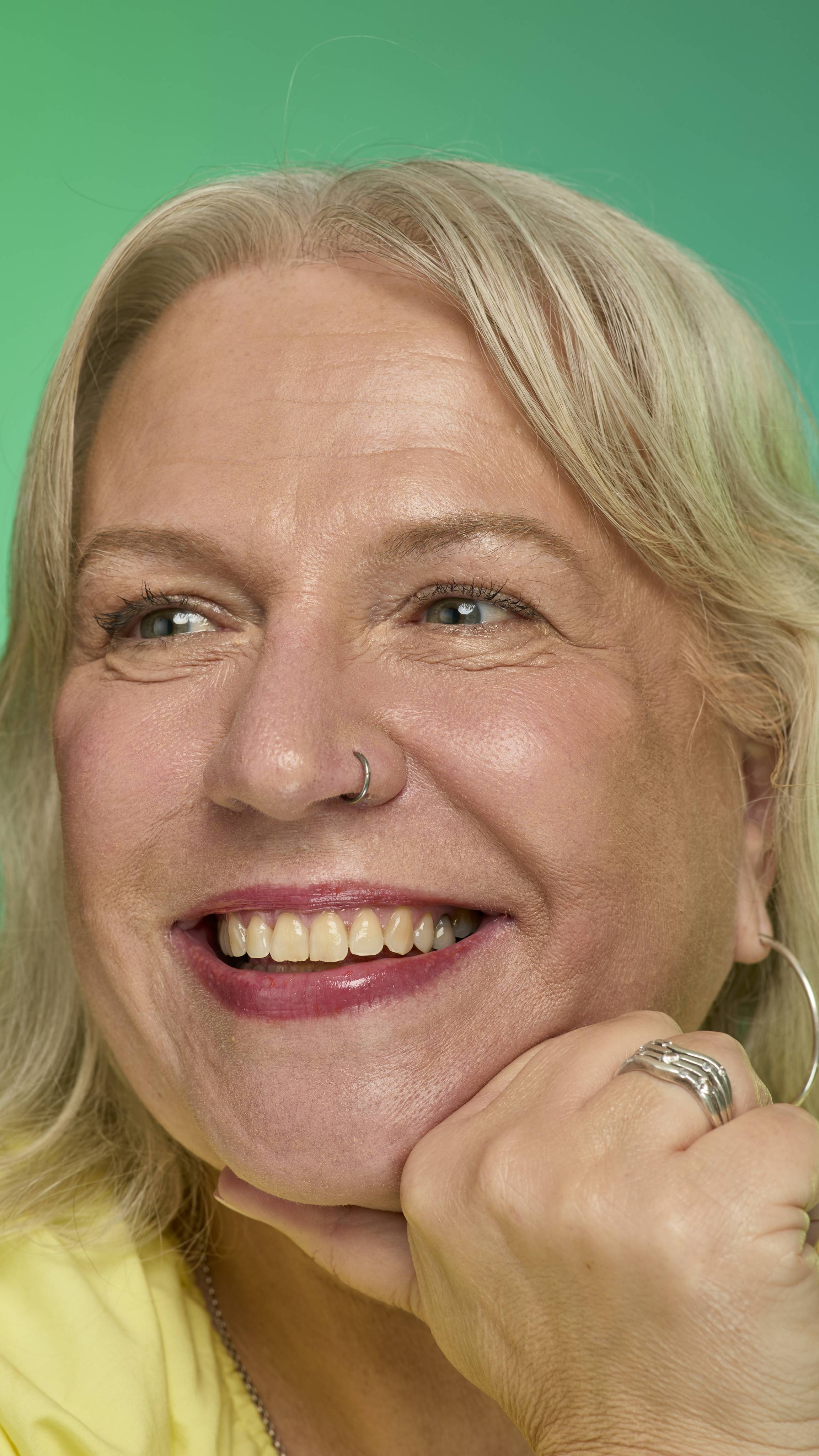 The image shows a model with blonde hair on a green and blue gradient background. They are smiling at the camera as their eyebrows look thick and full thanks to the Blonde brow pomade.