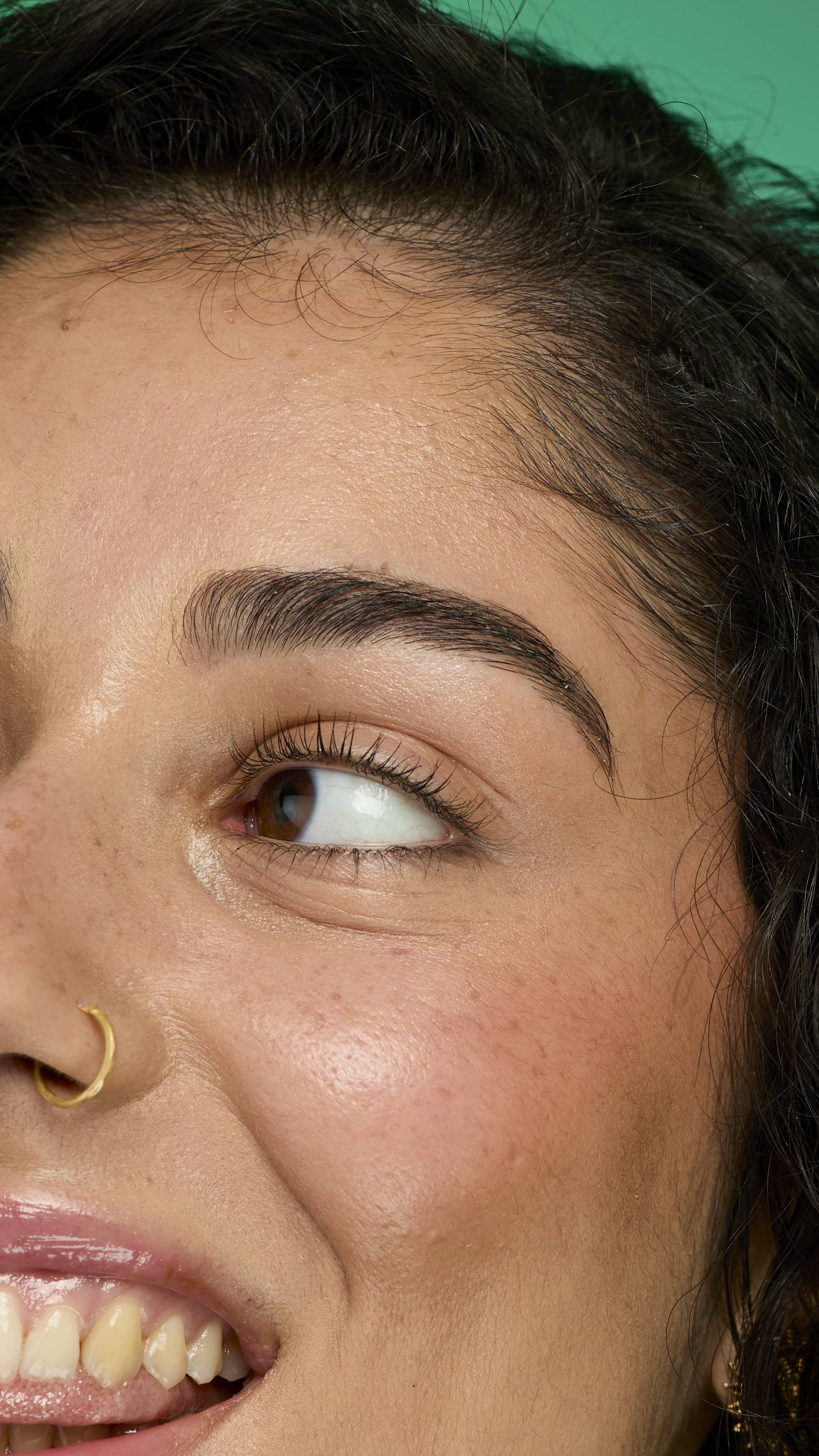 A close-up image of a model's face, with dark curly hair on a green background. They are smiling as they glance away. Their eyebrows look thick and full thanks to the Brun brow pomade.