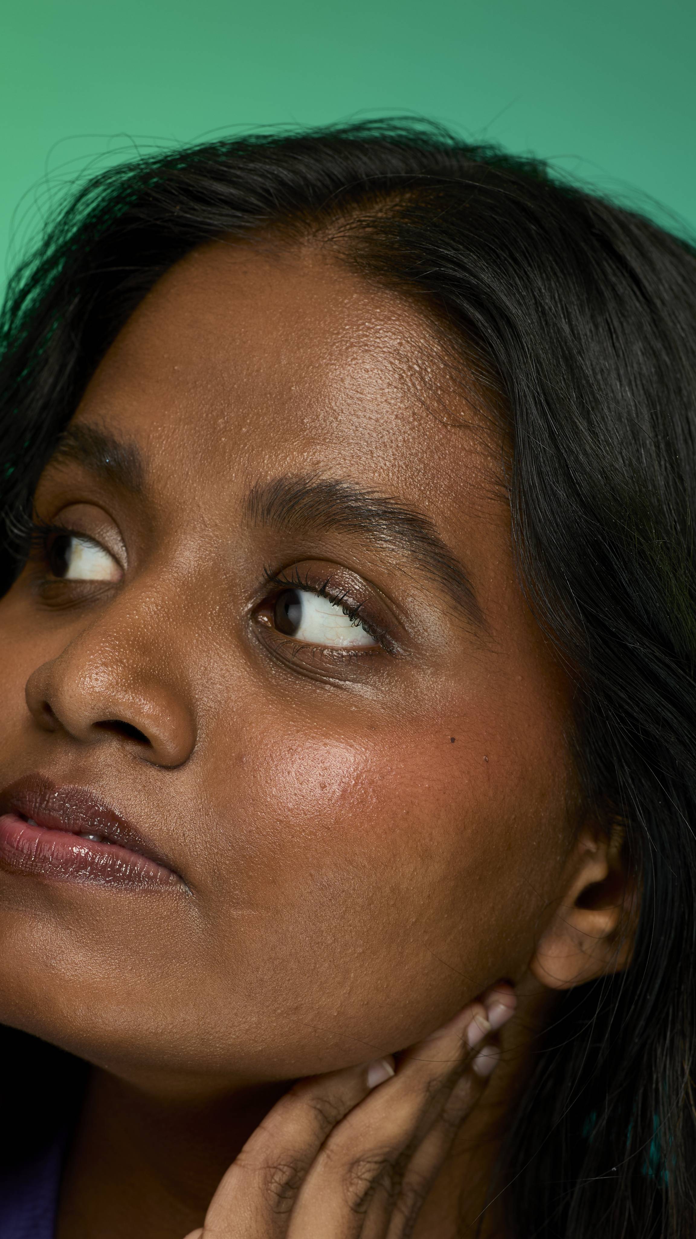 A close-up image of a model with dark straight hair on a green background. They are glancing away as their eyebrows look thick and full thanks to the Noir brow pomade.