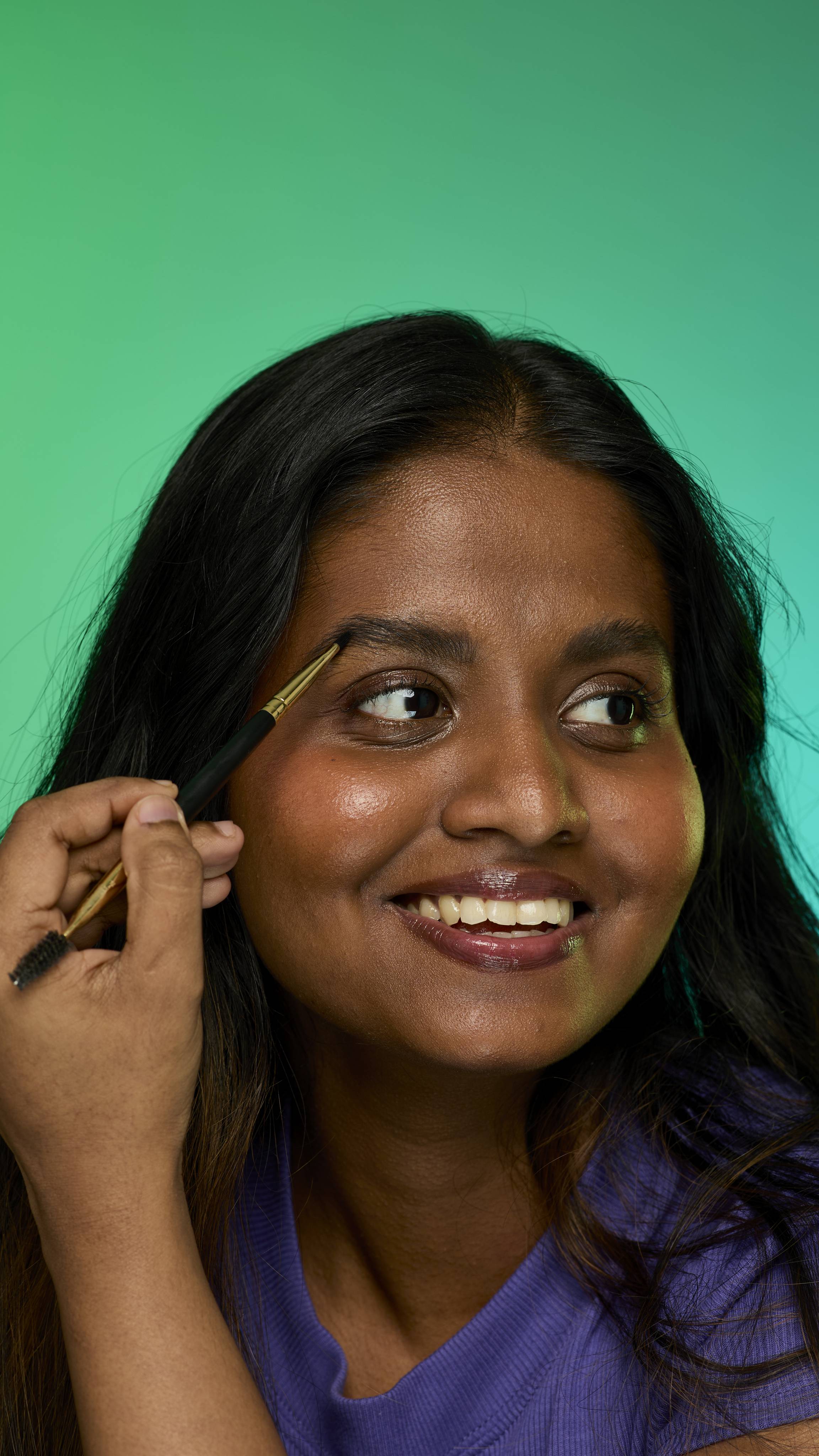 The image shows a model with dark straight hair on a green background. They are smiling and holding an eyebrow brush up to their face. Their eyebrows look thick and full thanks to the Noir brow pomade.