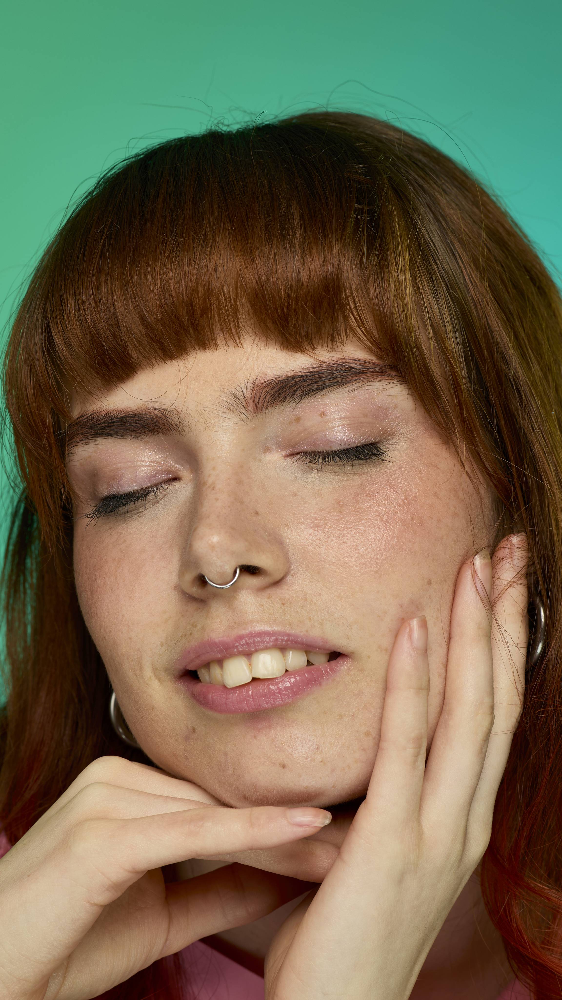 The image shows a model with auburn straight hair with a full fringe on a green and blue gradient background. They are closing their eyes as their eyebrows look thick and full thanks to the Rouge brow pomade.