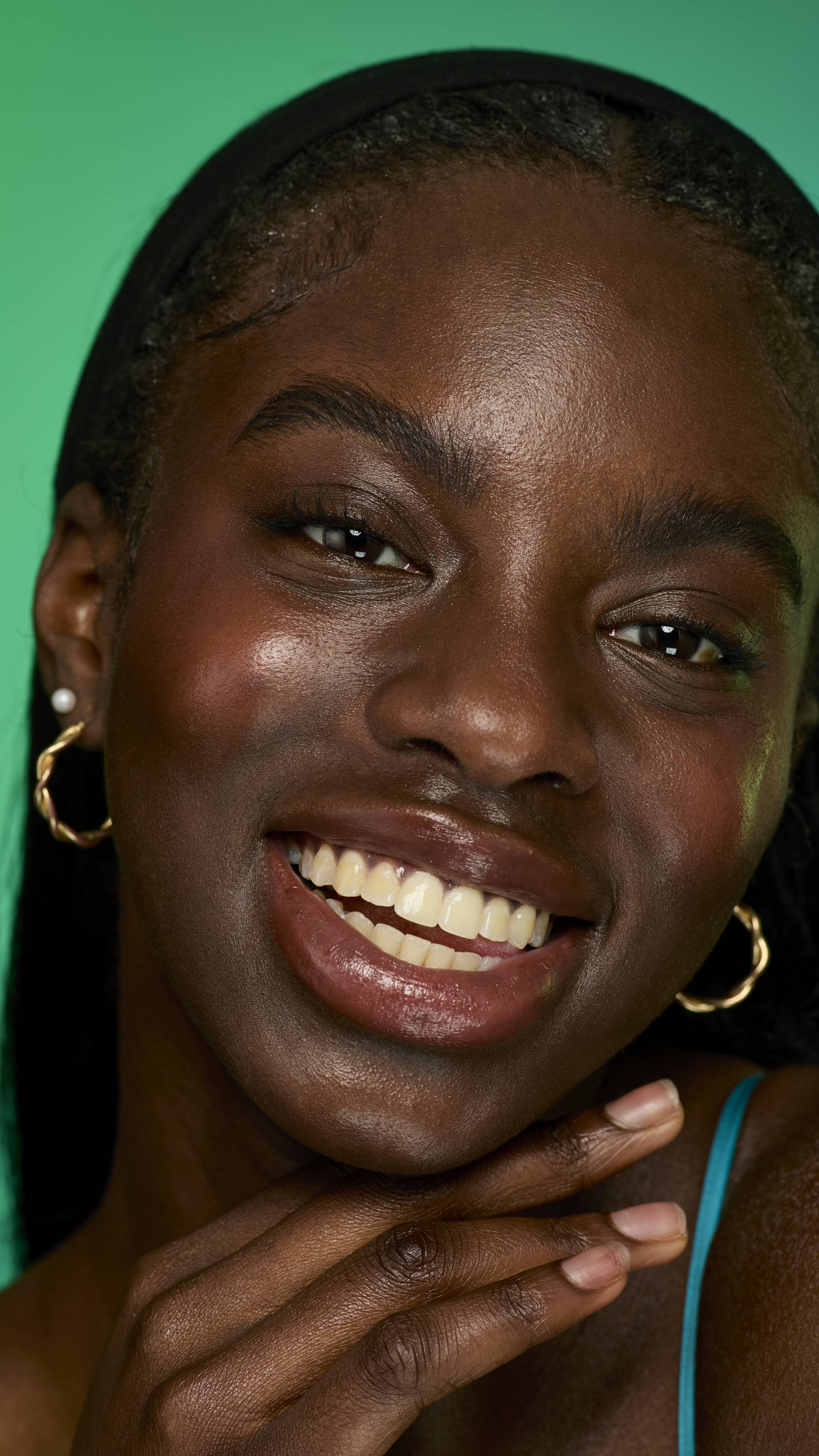 A close-up image of a model with dark straight hair on a green background. They are smiling with their hand at their face as their eyebrows look thick and full thanks to the Smoke brow pomade.