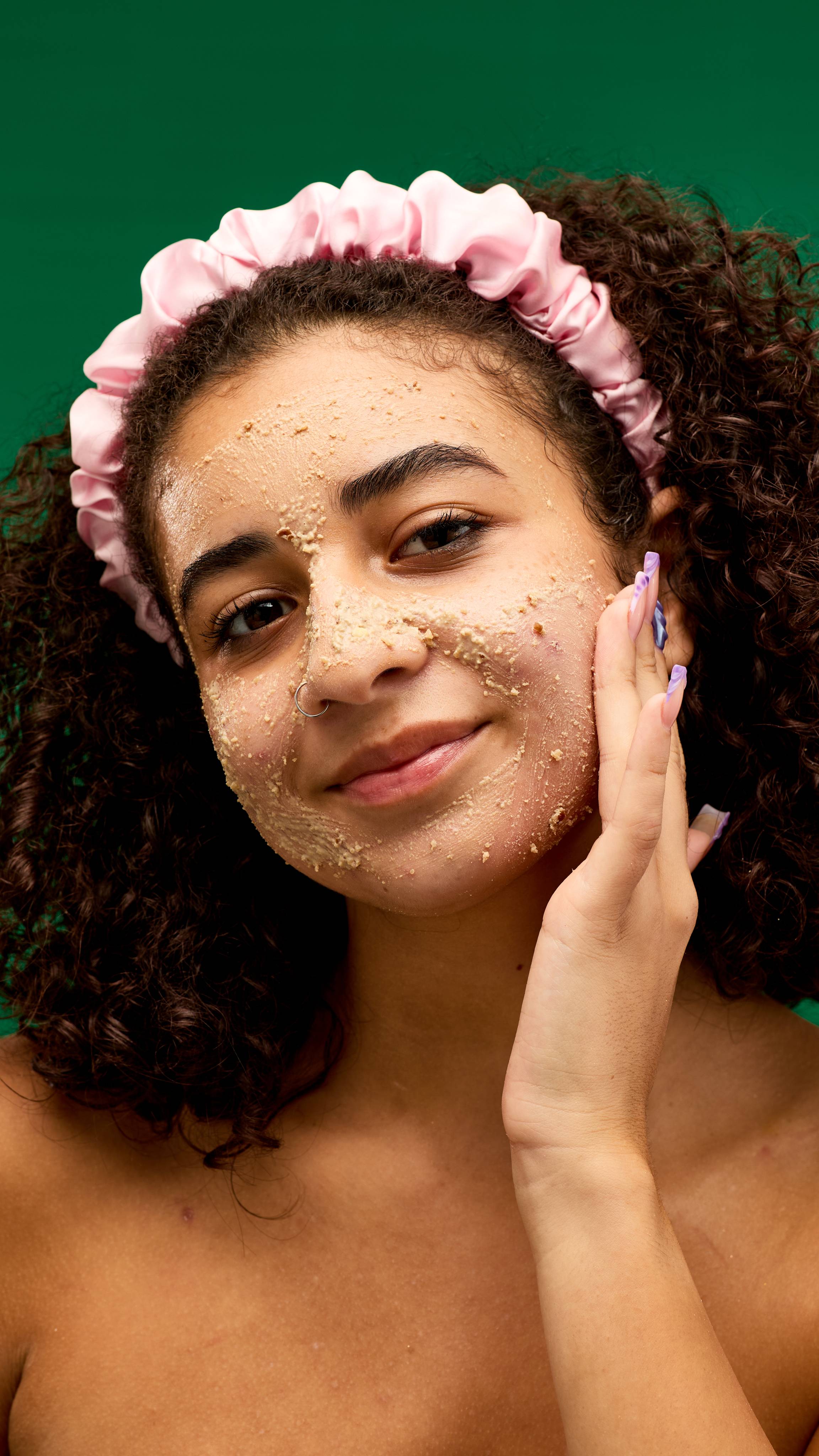 A person wearing a pink satin headband smiles while applying a LUSH Bûche de Noël cleanser to their face. The chunky, light-coloured cleanser is visible on their skin, and the background is a vibrant green.