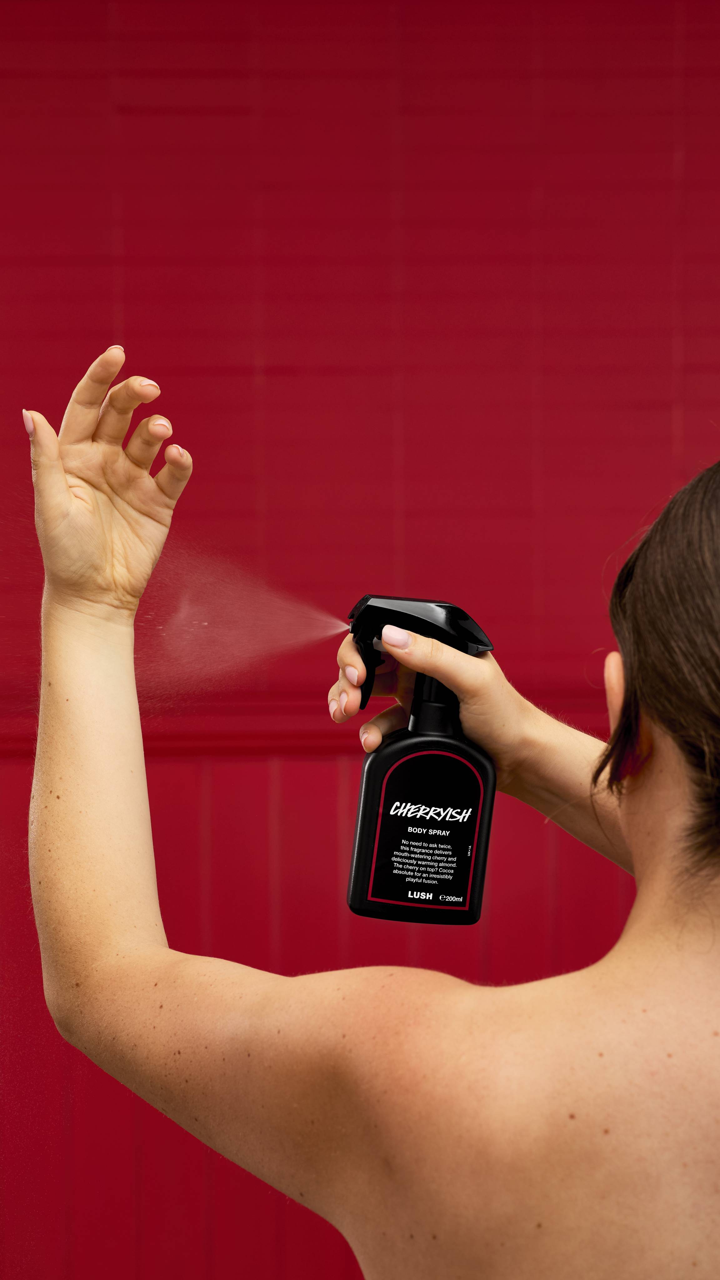 A recycled black LUSH body spray bottle with a trigger nozzle is held by a person from behind. The bottle, with its pink-rimmed "Cherryish" label, spritzes a mist into the air against a vibrant red background.