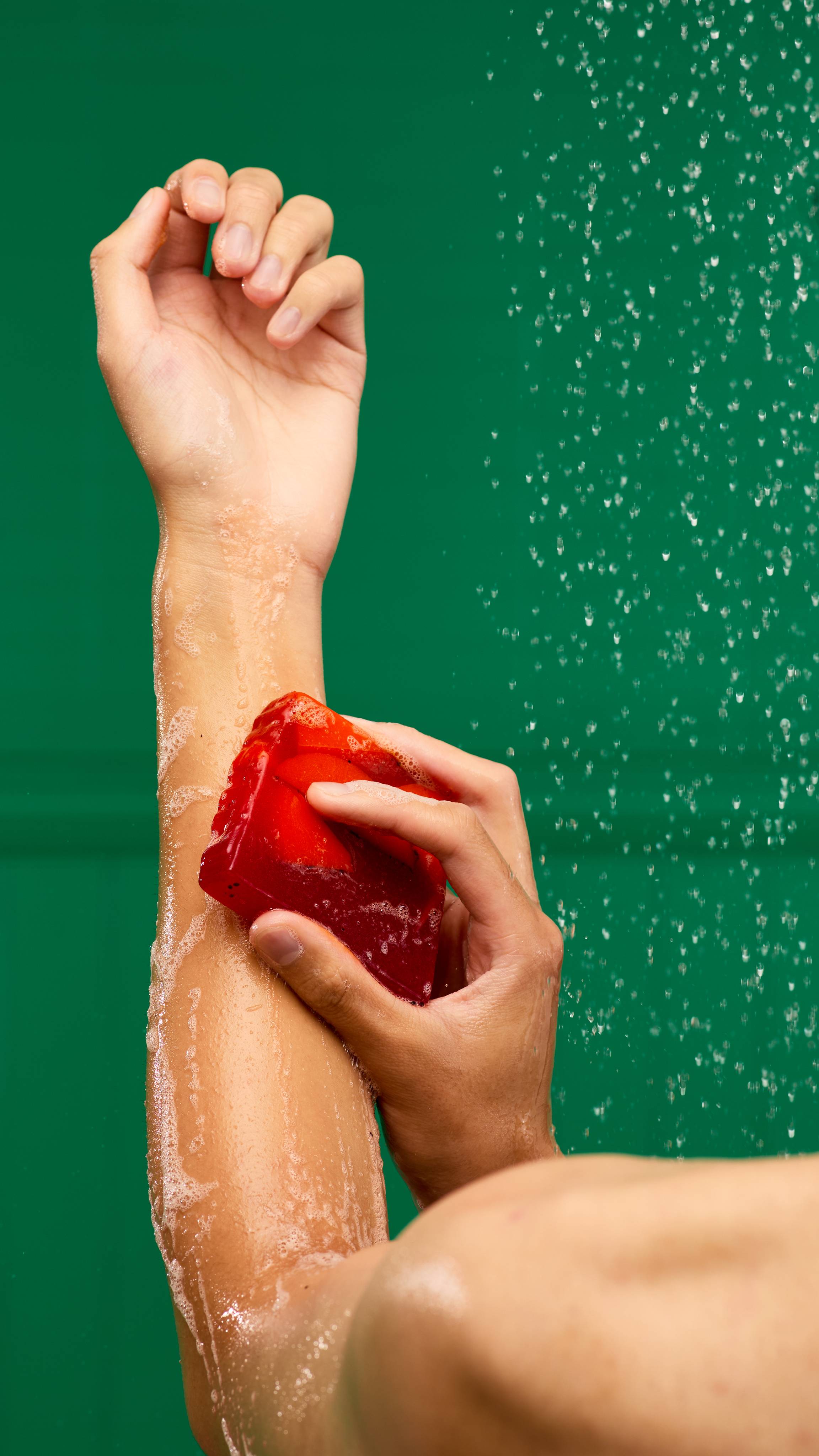 The image shows a light-toned arm reaching upwards in a shower. Water streams down as a slice of Christmas Cranberry soap is being used to wash their left forearm. The soap is creating a rich lather on a green background.