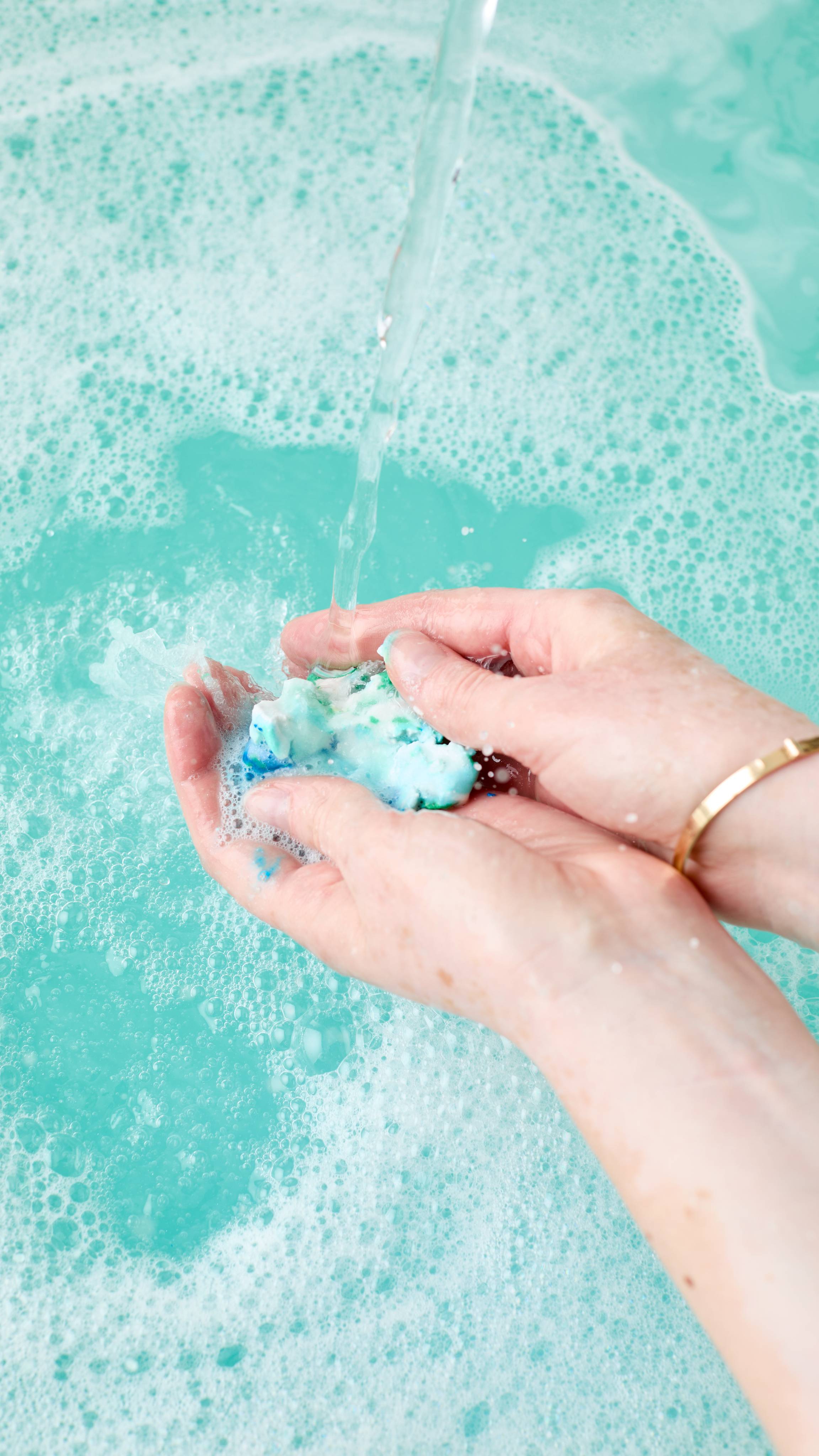 Two fair-skinned hands gently squeeze and crumble some of the Christmas Lights Fun product under a stream of water, creating teal-coloured water and white foam in a bathtub.