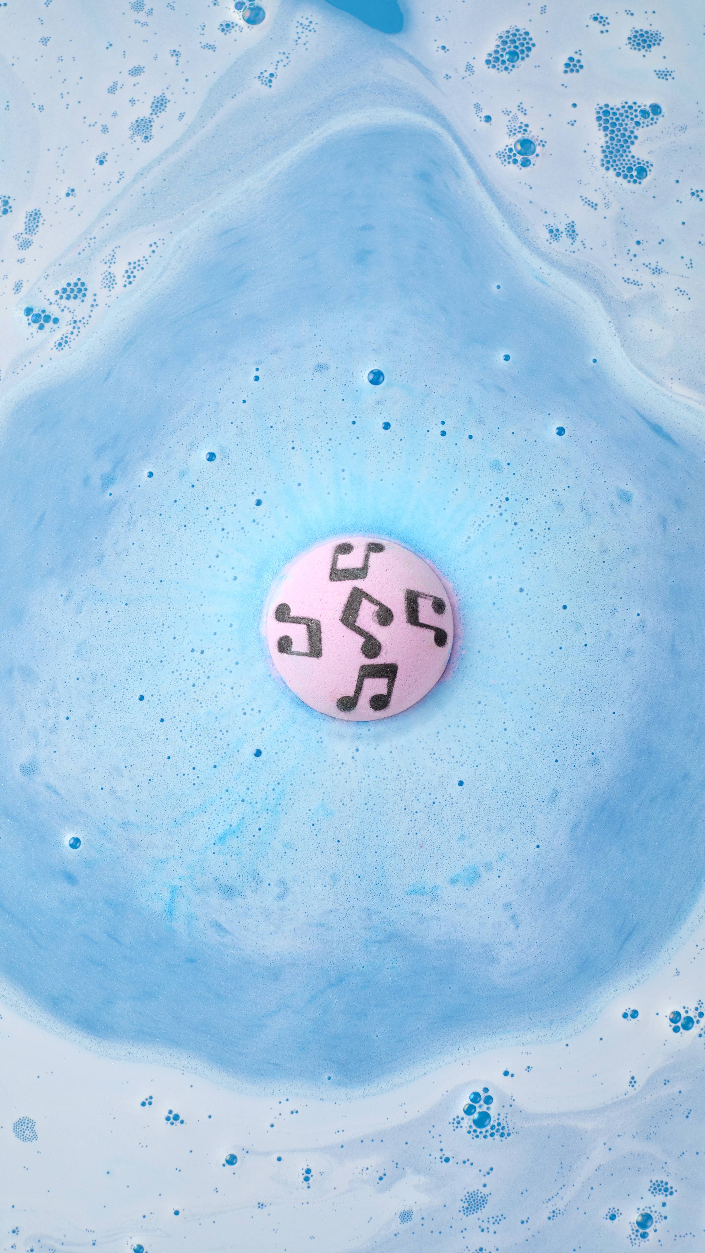 An aerial view of the "Dad Dancing" bath bomb with a pink top and black musical notes, dissolving in a bath. It creates a soft blue circle of fizz on the water's surface, surrounded by white soapy foam and scattered blue bubbles.