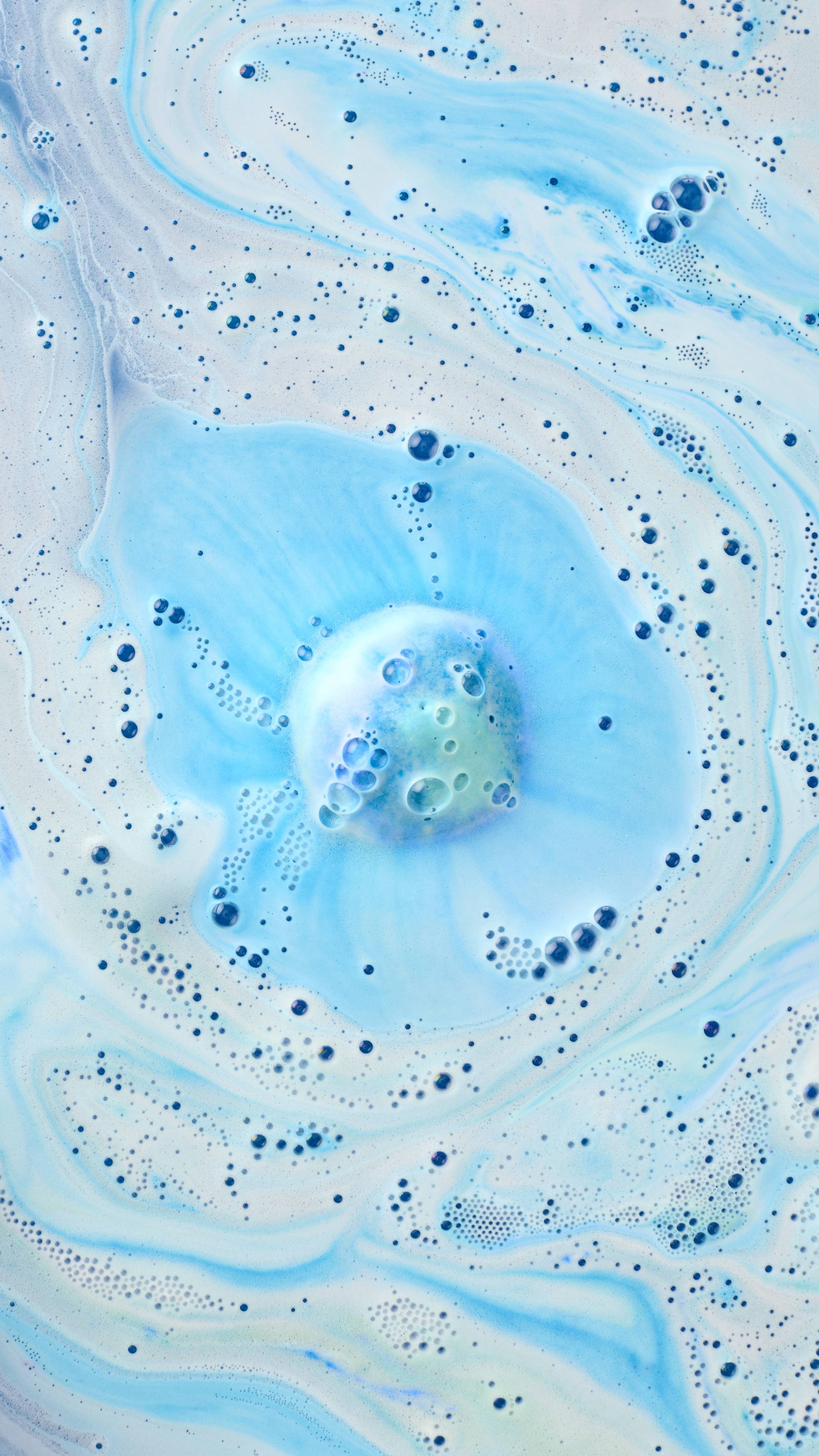 An aerial view of a bathtub filled with pale blue water and swirling white foam. In the centre, a partially dissolved "Easy Breezy" bath bomb creates a concentrated pool of bright blue fizz and clusters of small bubbles as it releases colour.