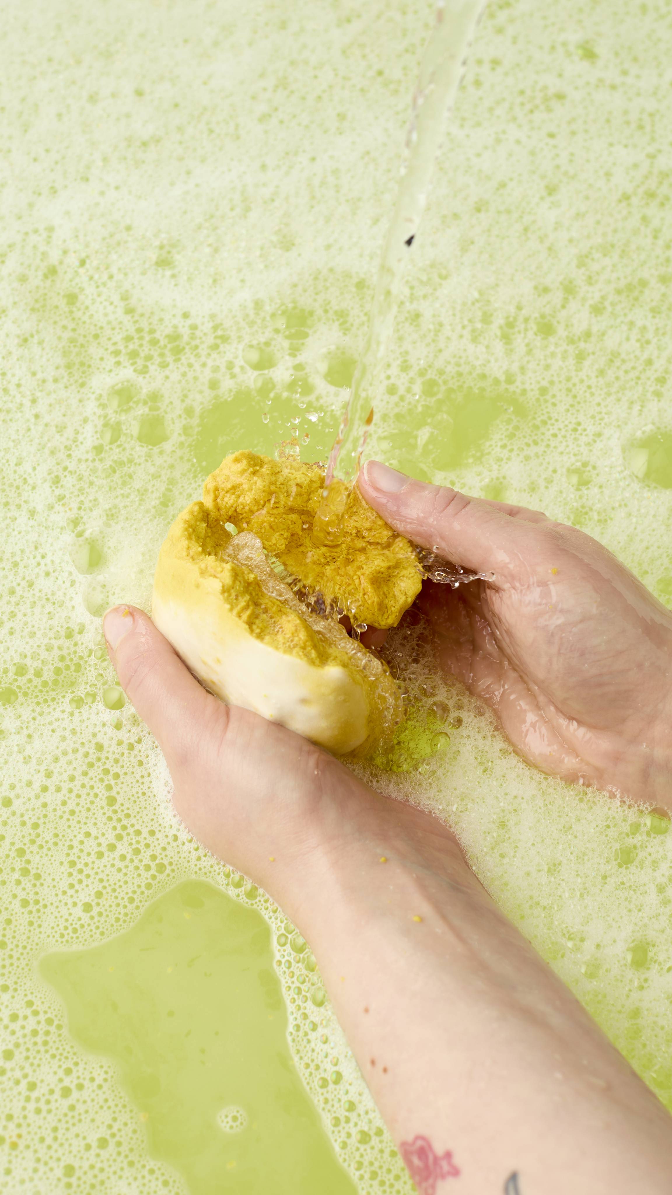 A model breaks the "Easy Peasy Lemon Squeezy" bubble bar in half under a running tap. The bubble bar crumbles, creating a frothy white lather that blends into the surrounding pale yellow, bubbly bathwater.