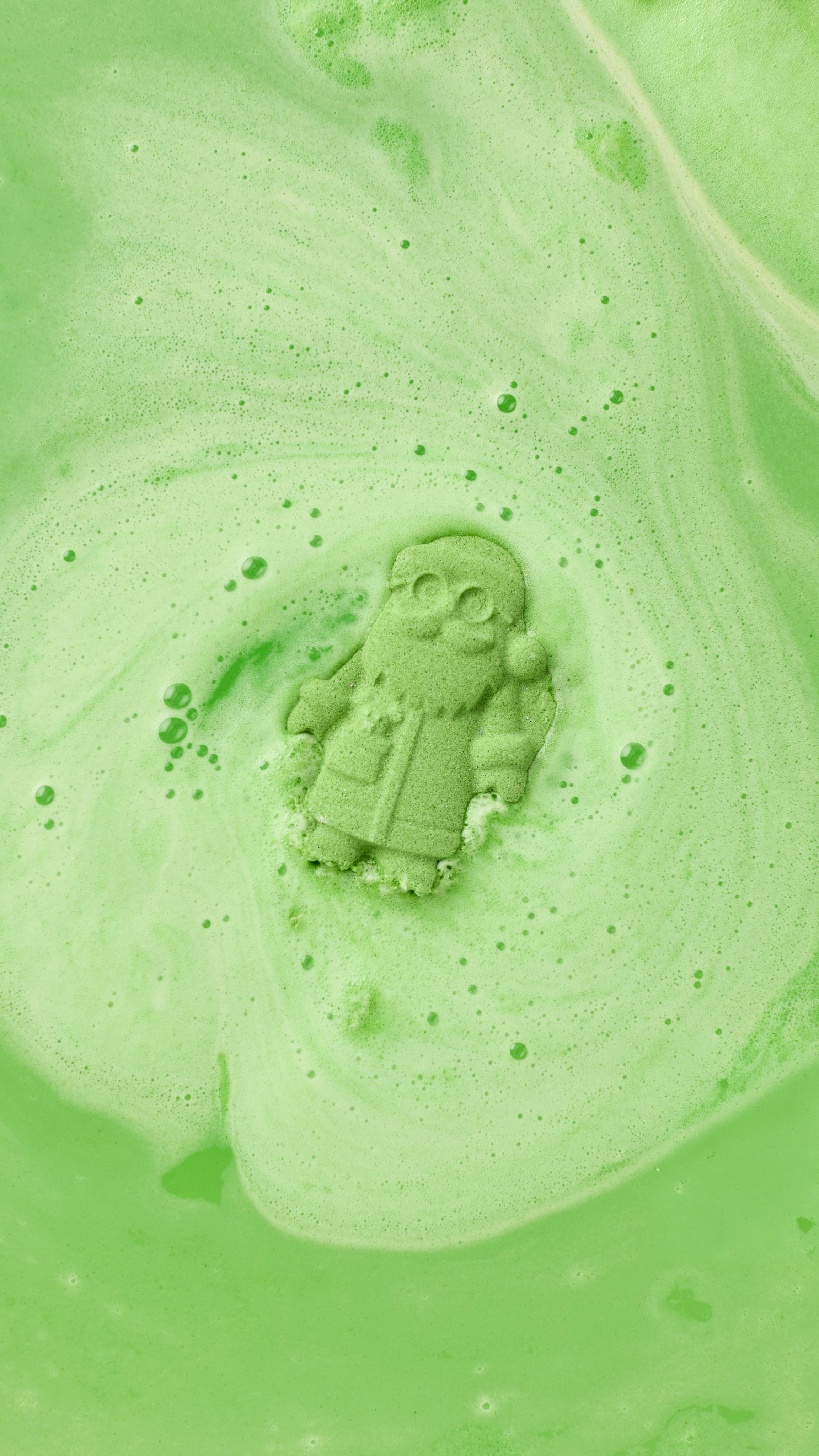 The image shows a speckled green Father Christmas bath bomb fizzing in bright green bath water. Thick green foam and bubbles swirl outwards, almost obscuring the Santa-shaped product as it dissolves.