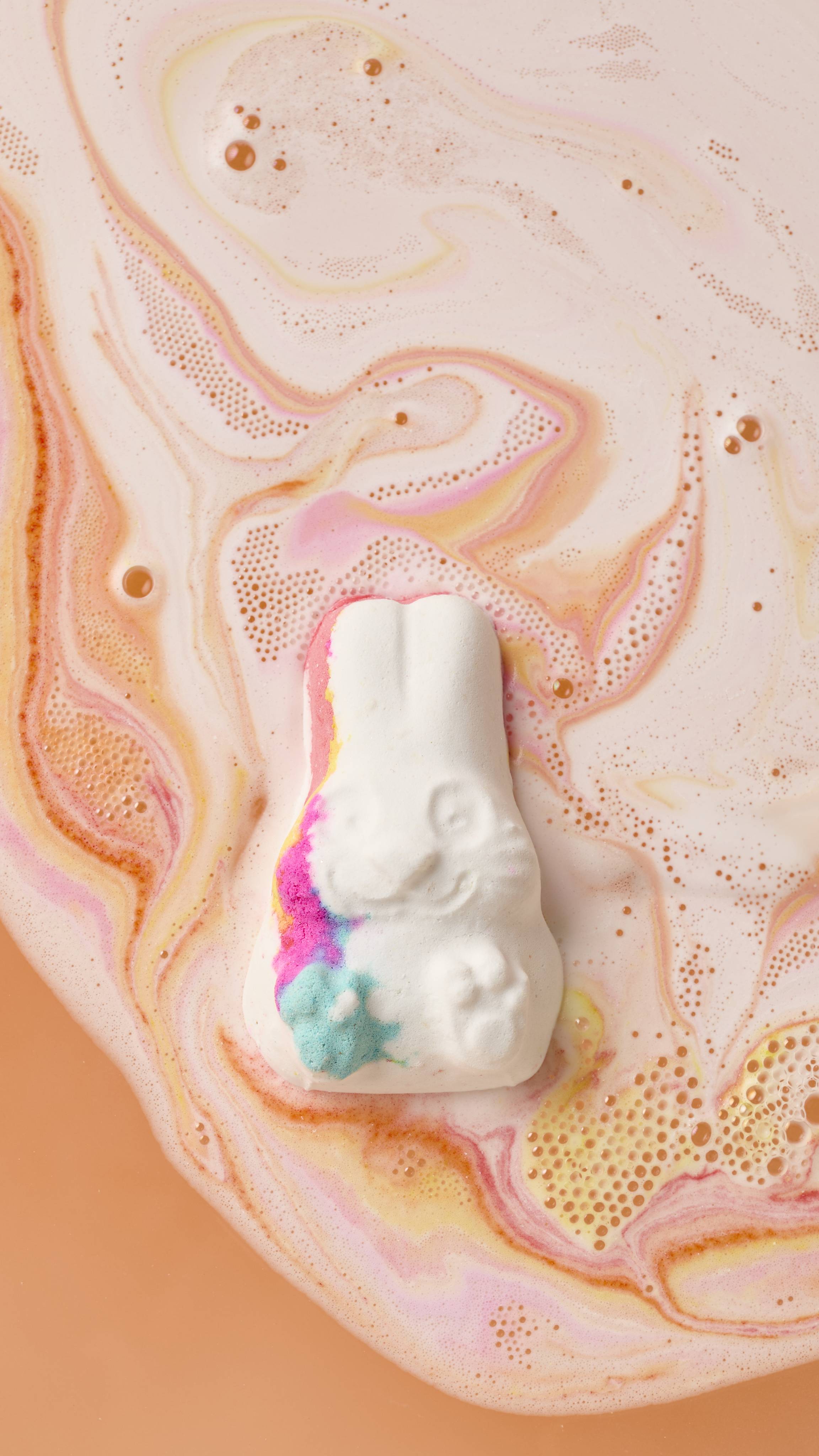 A white, rabbit-shaped Follow The White Rabbit bath bomb floats in peach-colored water. It features vibrant pink, blue, and yellow streaks as it dissolves, creating colorful swirls.