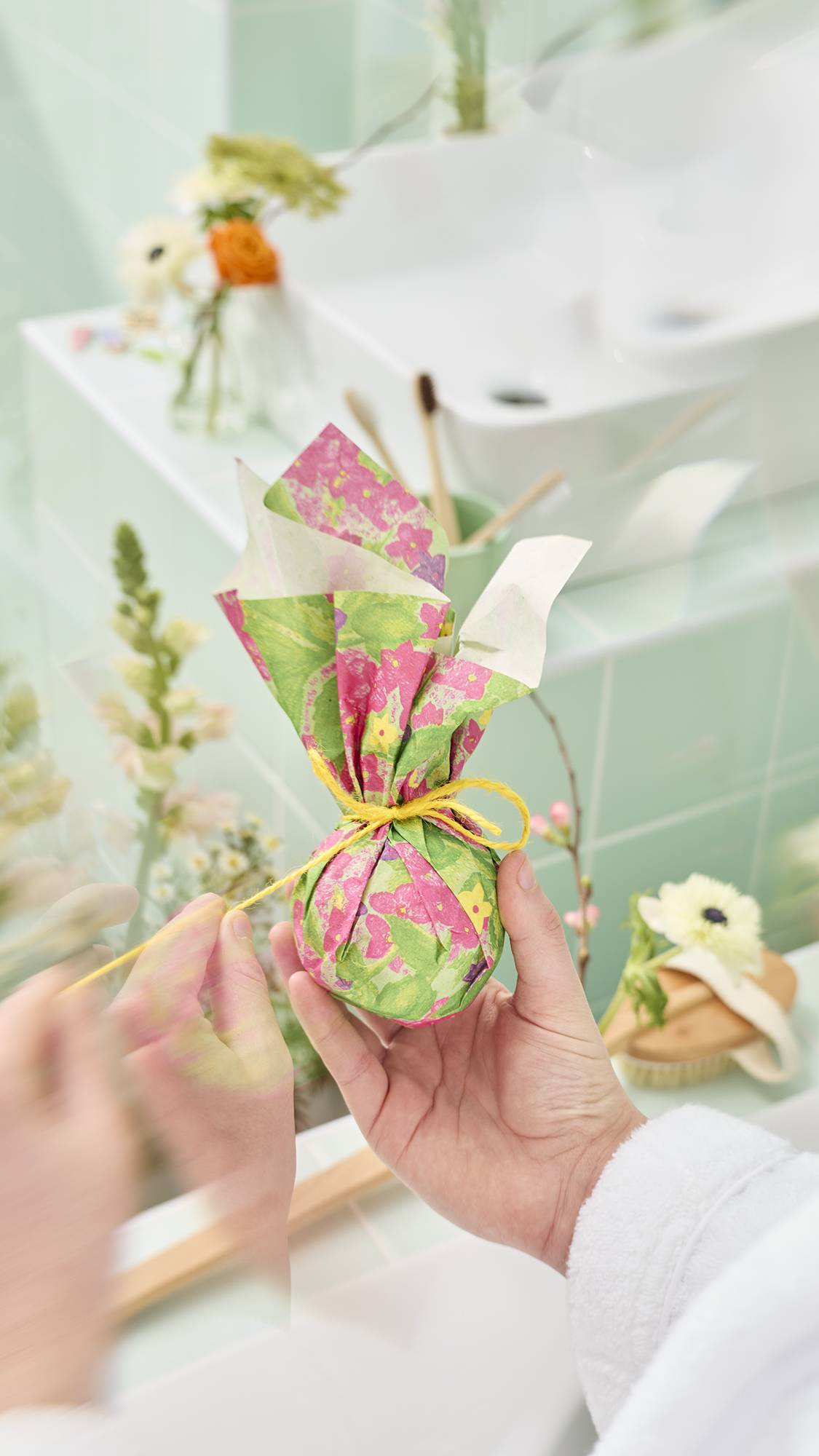 The image shows a colourful sheet of "Garden in Spring" Lokta paper featuring a hand-painted floral design, wrapped around a spherical bath bomb gift and tied with a yellow twine bow. The background features a tiled background.